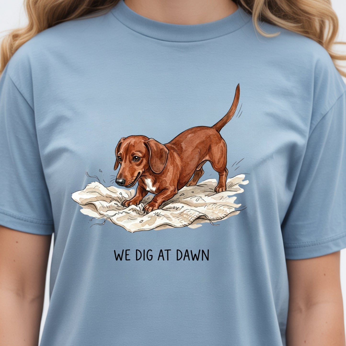 A person wearing a light blue crewneck t-shirt with a graphic of a brown dachshund digging in snow and the text 'WE DIG AT DAWN' printed below.