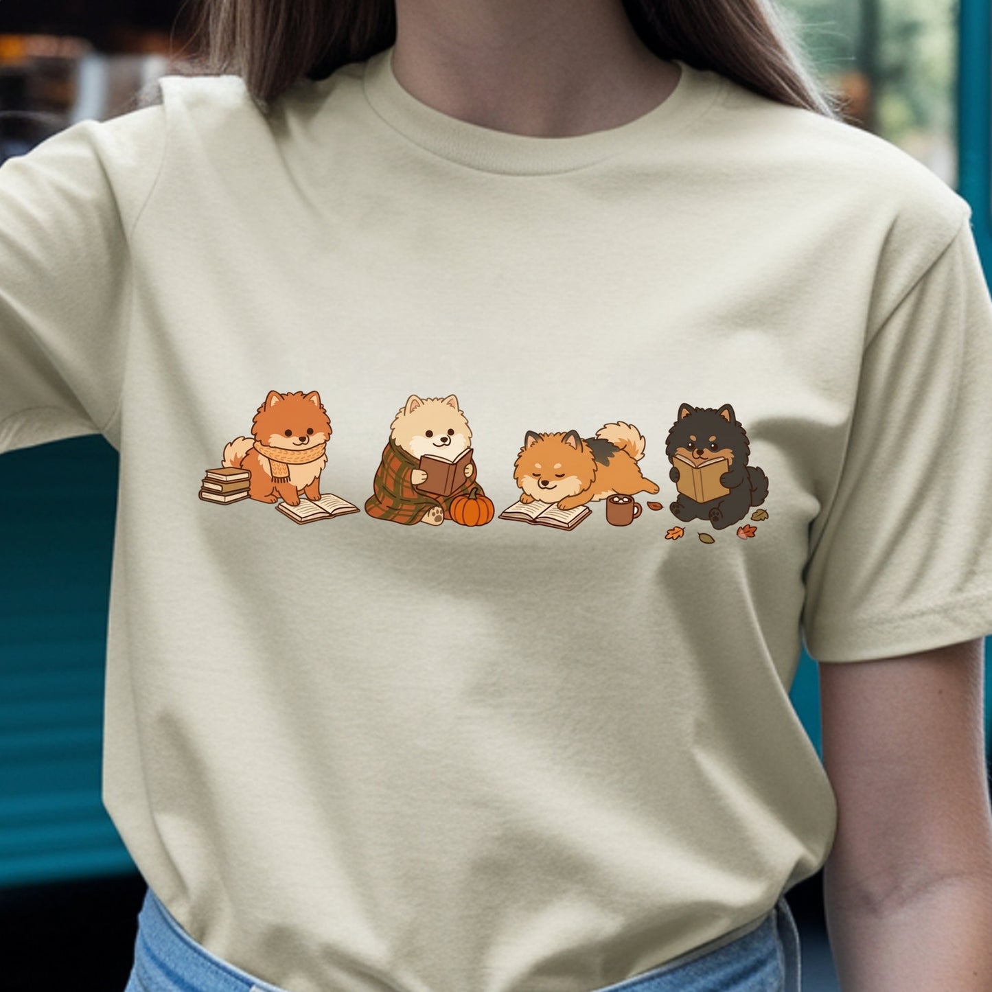 A person wearing a light beige crewneck t-shirt with a graphic print of four cute cartoon Pomeranian dogs reading books in an autumn theme, paired with blue jeans.