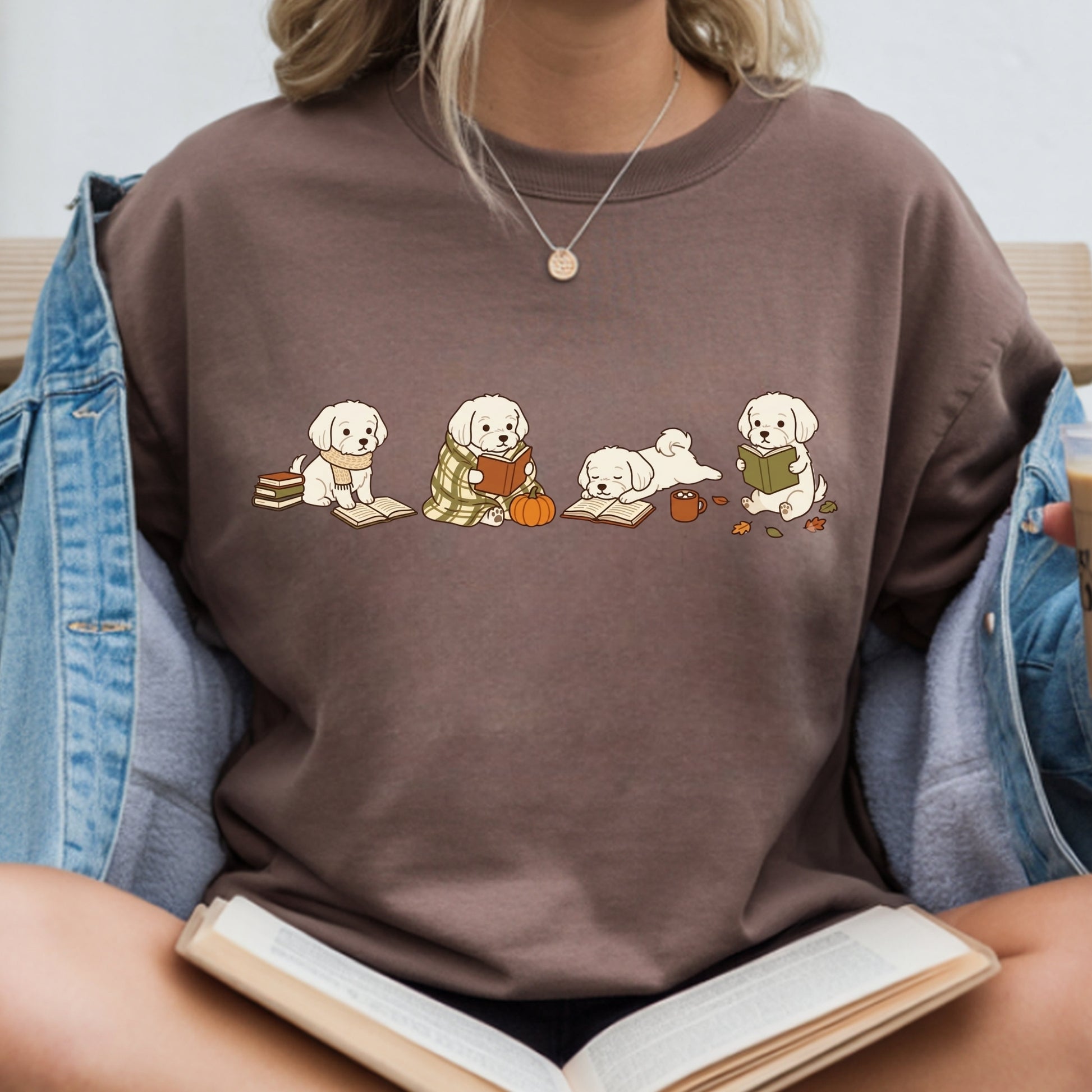 A person wearing a brown crewneck sweatshirt with a graphic of four white puppies reading books in an autumn setting. The person is also wearing a light blue denim jacket and holding an open book.