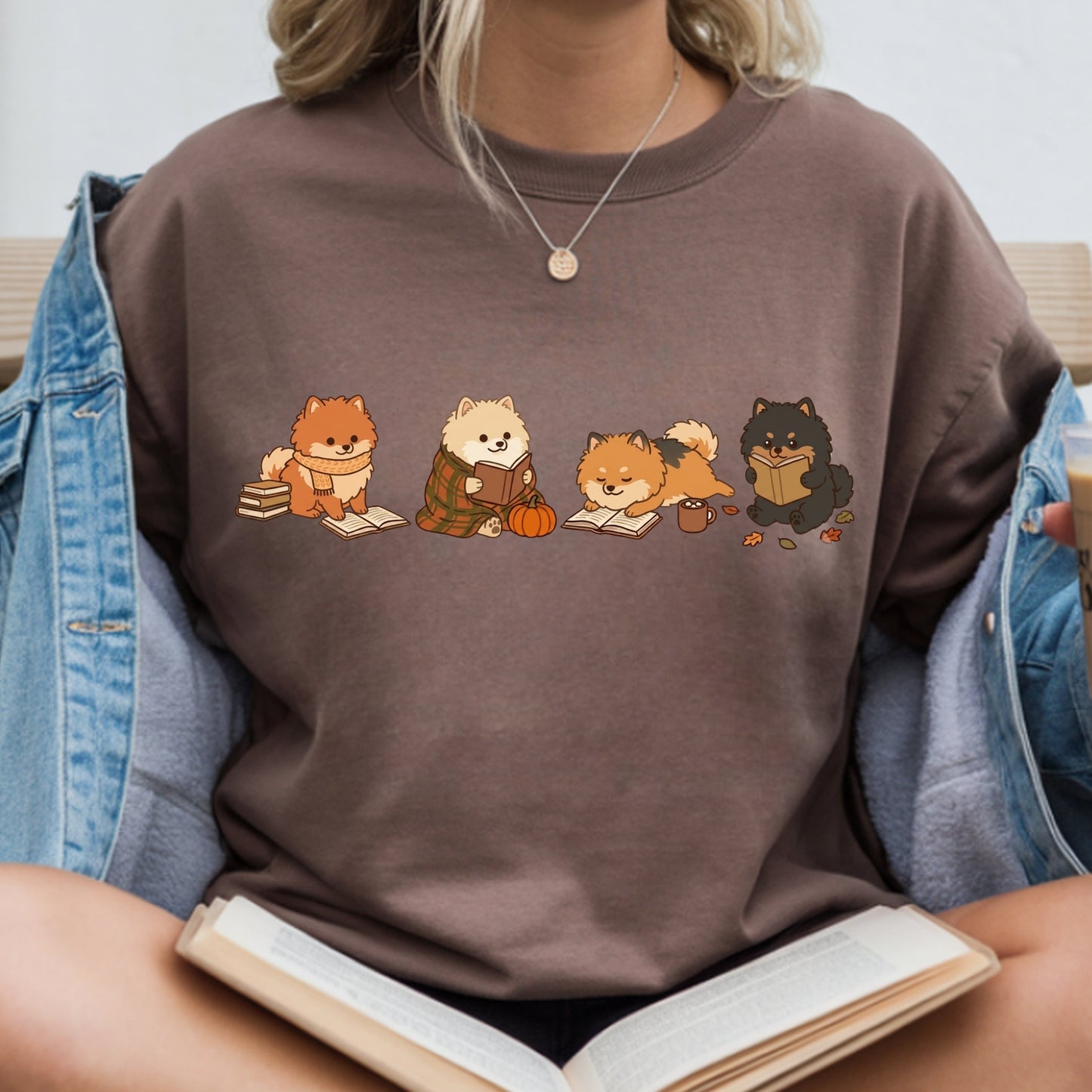 A person wearing a brown crewneck sweatshirt with a graphic of four cute dogs reading books, styled with an open denim jacket and holding an open book.