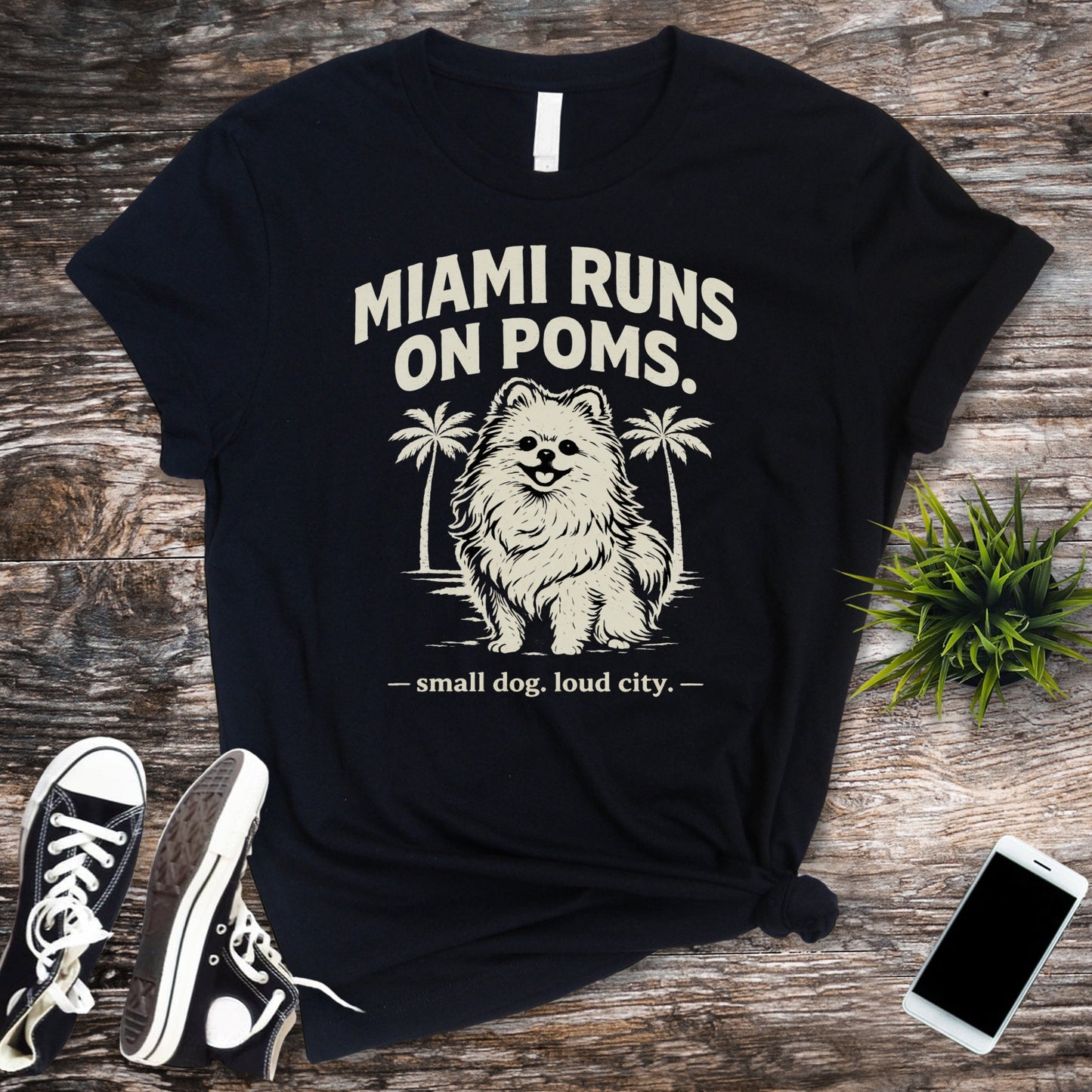 Black t-shirt with a dog graphic and text on a wooden surface with sneakers and a phone.