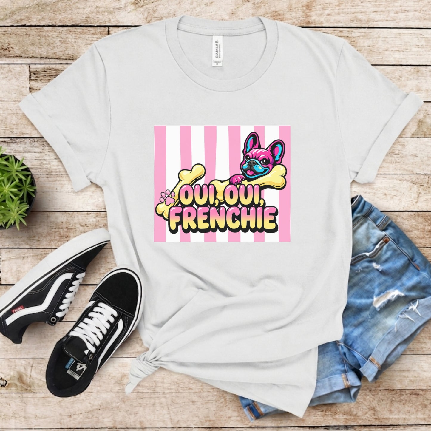 White t-shirt with colorful graphic design on a wooden surface with sneakers and jeans.