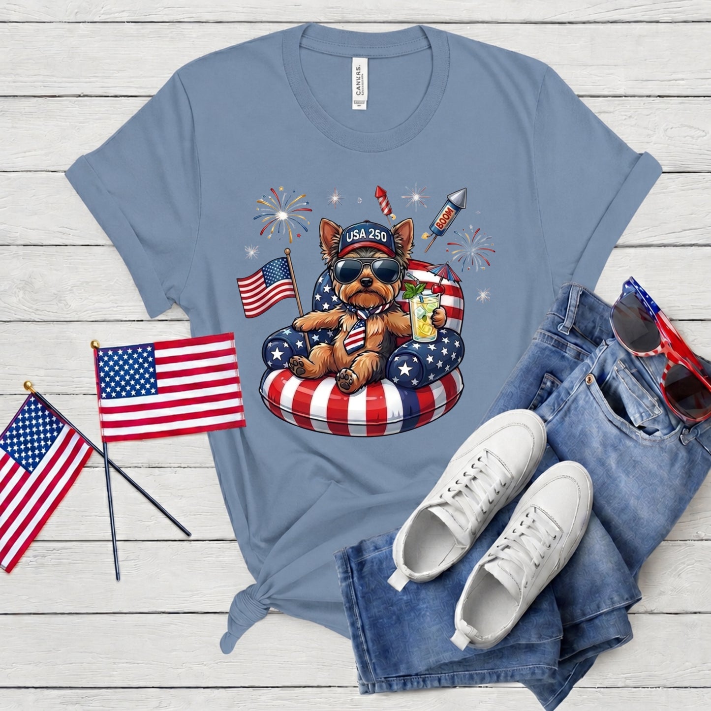 Blue t-shirt with a dog graphic, American flags, and sunglasses on a wooden surface.