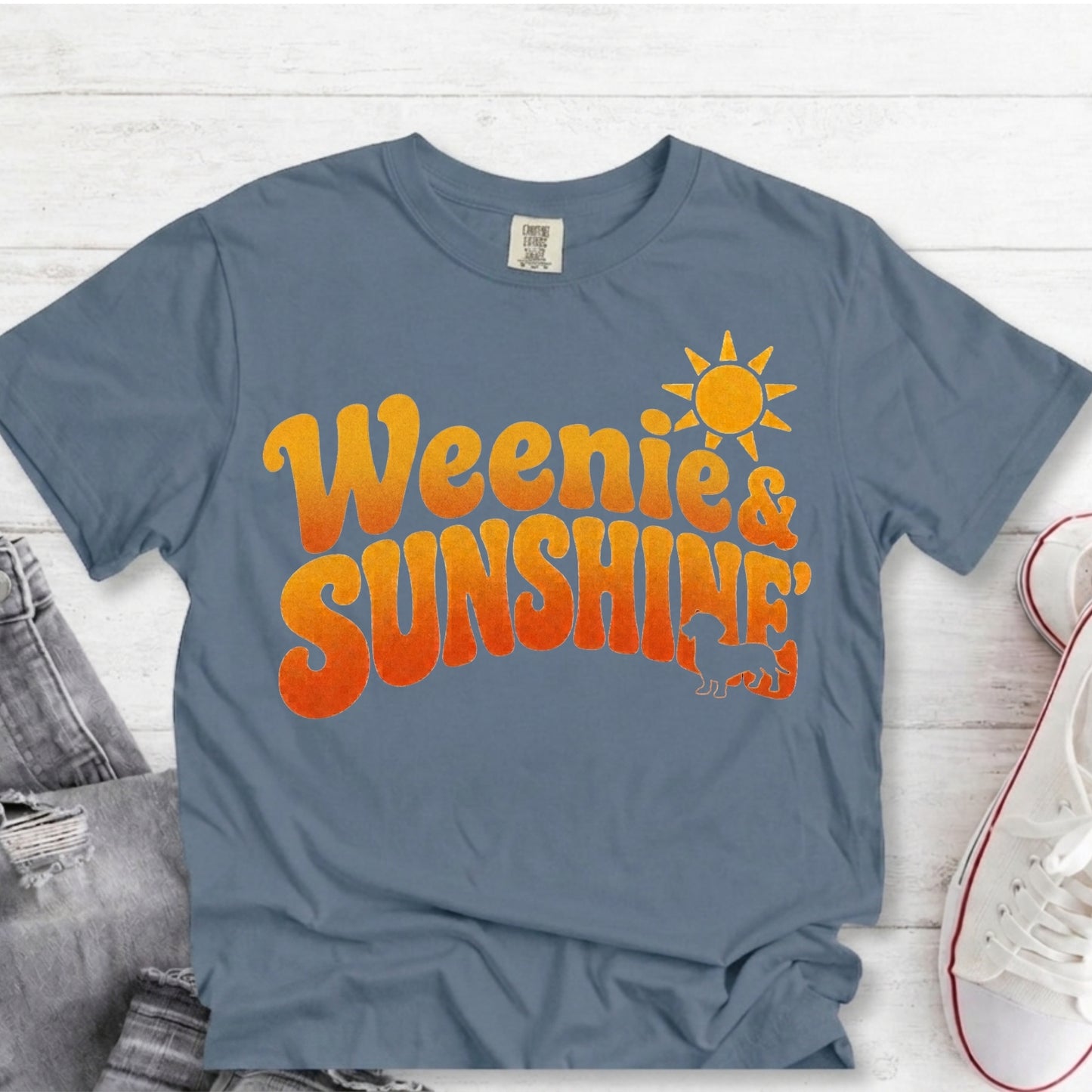 A slate blue graphic t-shirt laid flat on a white wood background, styled with a pair of gray jeans and white sneakers. The t-shirt features a retro-style orange and yellow gradient text graphic reading 'Weenie & SUNSHINE' with a yellow sun and small dinosaur graphic.
