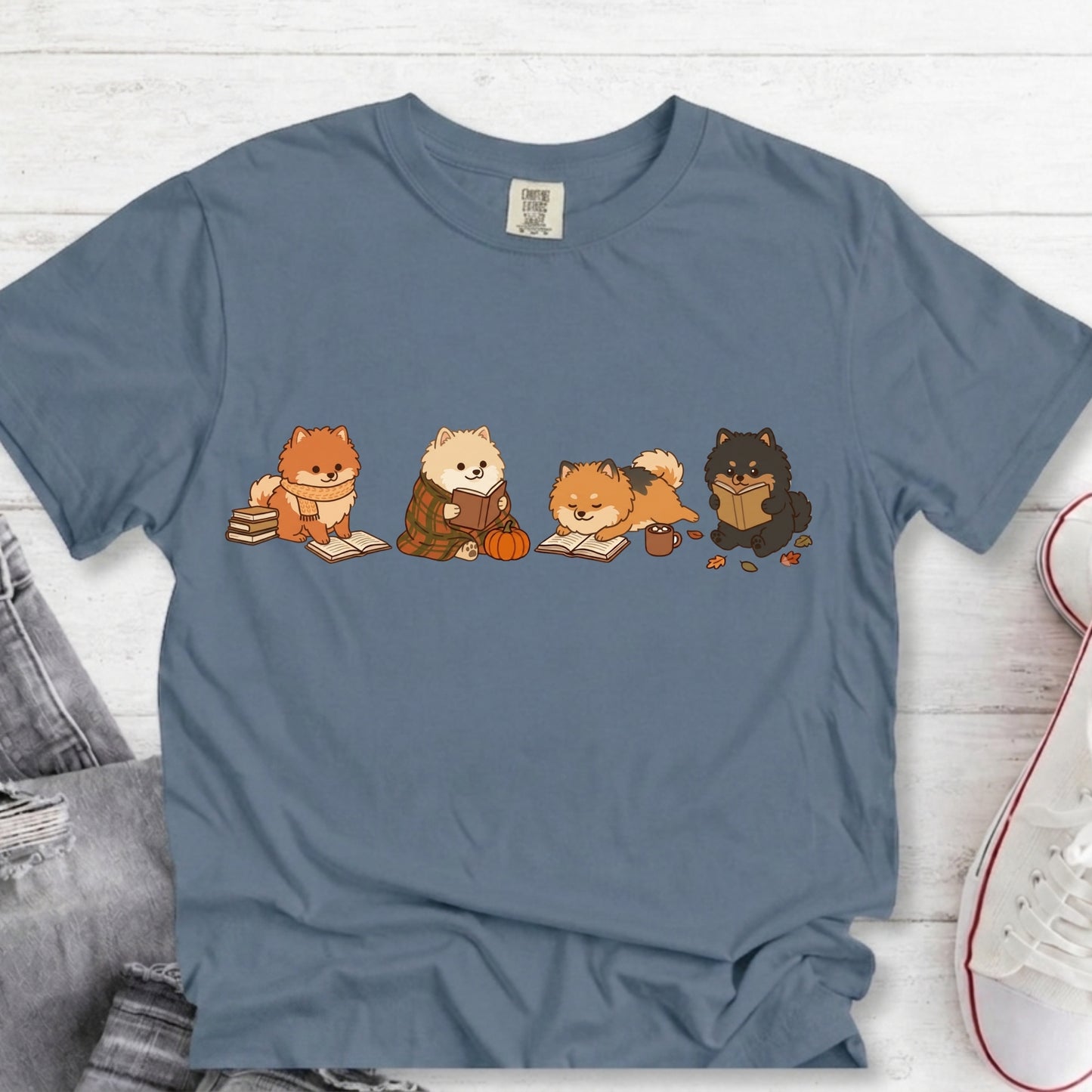 A slate blue crewneck t-shirt featuring a graphic of four cute cartoon dogs reading books, styled in a flat-lay on a white wood background with jeans and a sneaker partially visible.