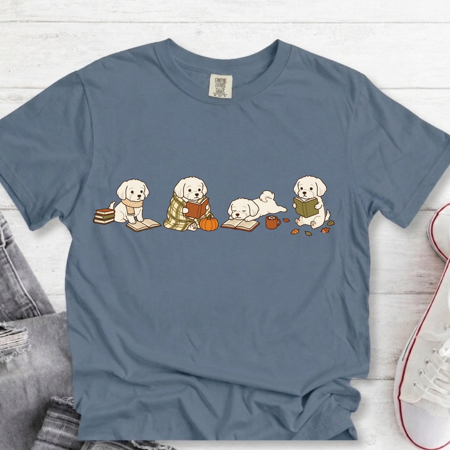 A slate blue crewneck t-shirt featuring a graphic of four cute puppy characters reading books, styled in a flat-lay on a white wood background with jeans and a sneaker partially visible.