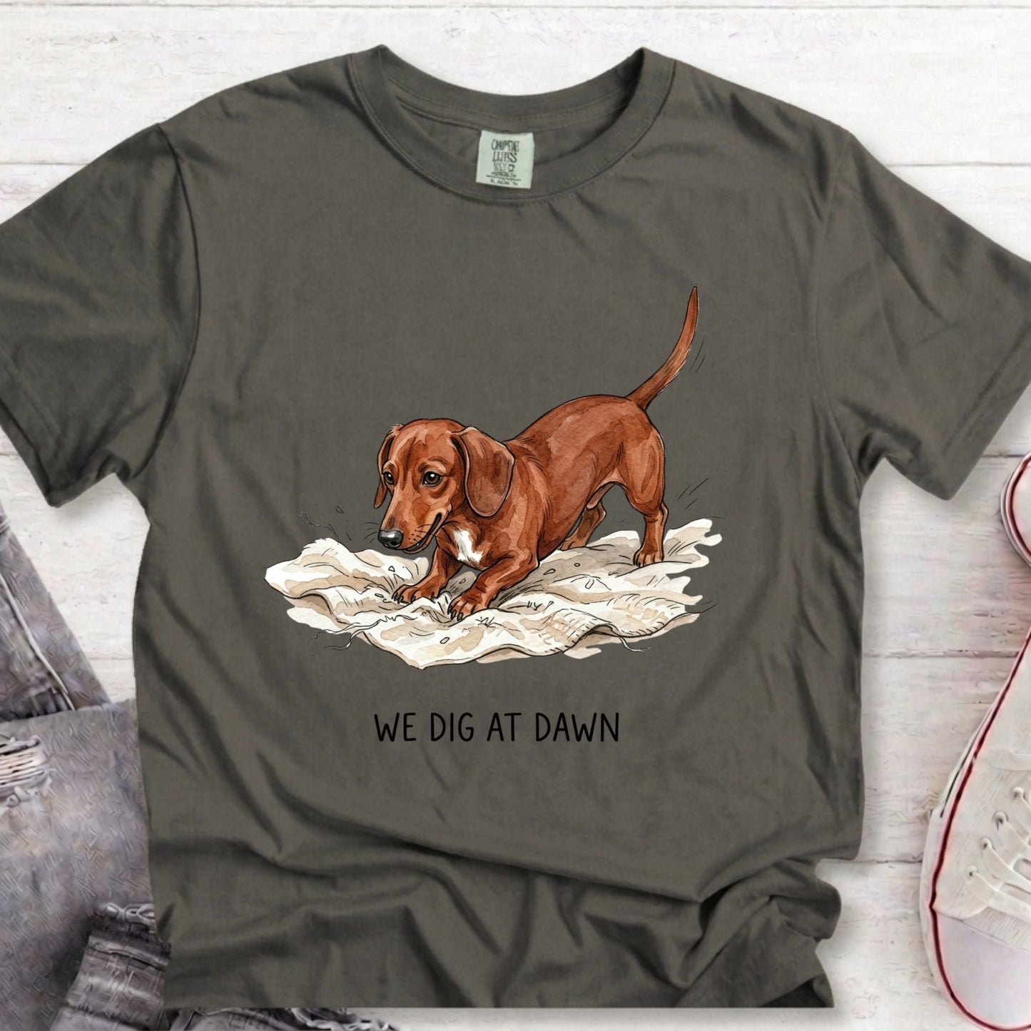 A dark gray t-shirt featuring a graphic of a brown dachshund digging in white soil with the text 'WE DIG AT DAWN' printed below. The t-shirt is laid flat on a white wood background with jeans and a sneaker partially visible as styling props.