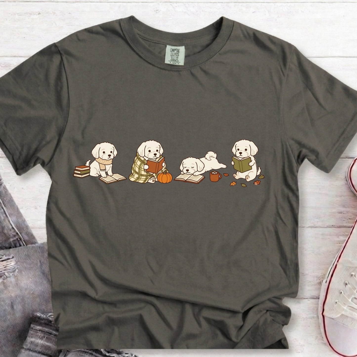 A dark gray crewneck t-shirt featuring a graphic of four cute puppy illustrations reading books, styled in a flat-lay on a white wood background with jeans and a sneaker partially visible.