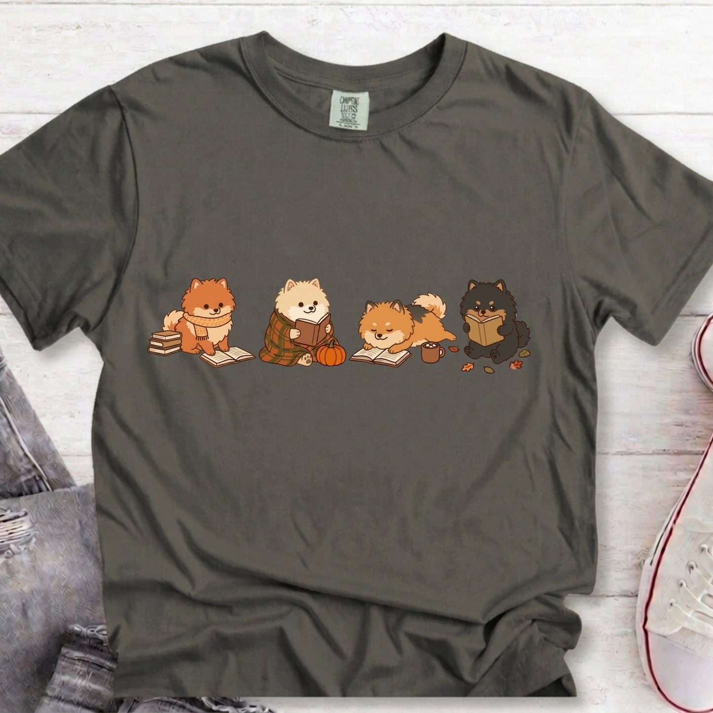 A dark gray crewneck t-shirt featuring a graphic of five cute cartoon dogs dressed in autumn clothing, laid flat on a white wood background with jeans and a sneaker partially visible.