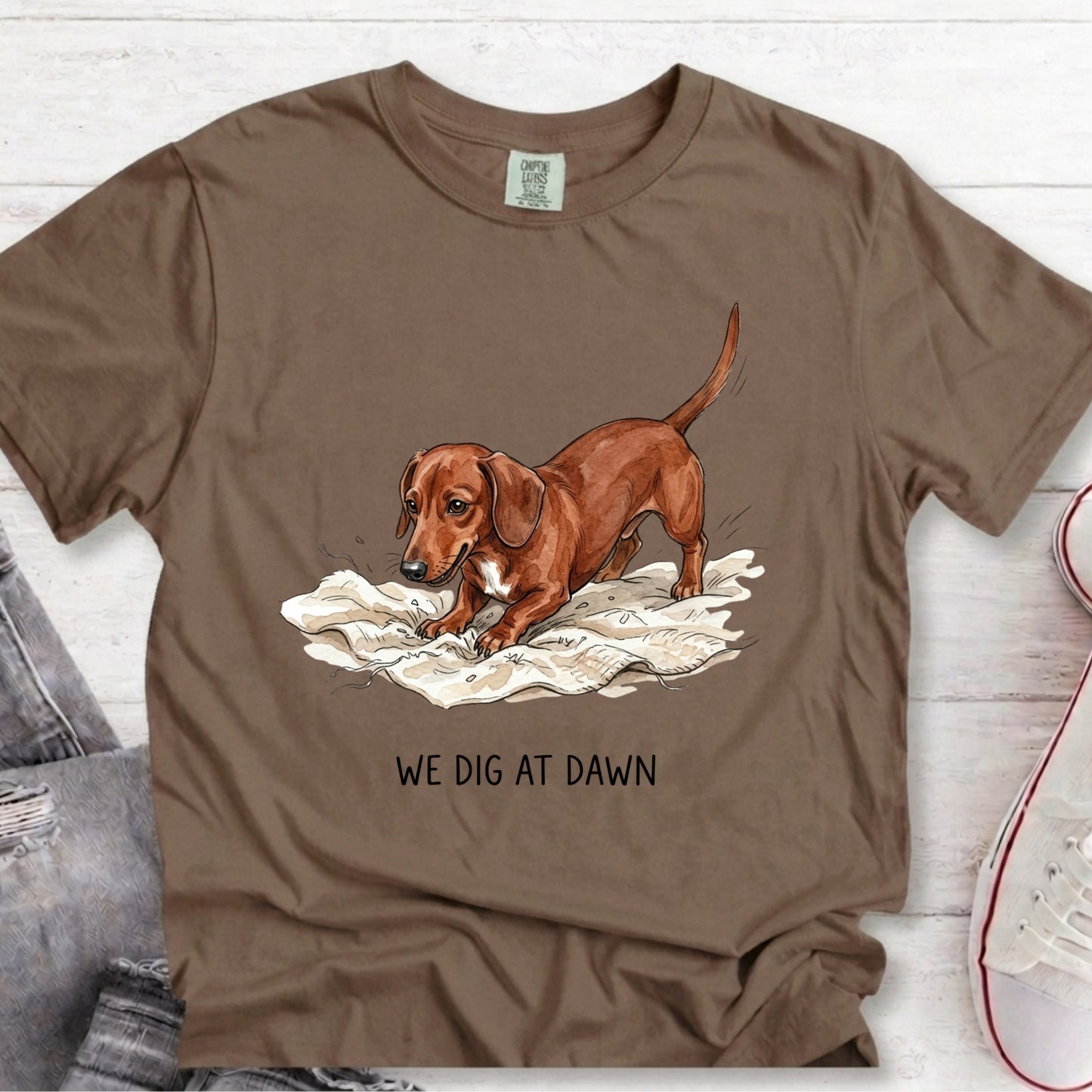 A brown short-sleeve graphic t-shirt laid flat on a white wood background, featuring an illustration of a dachshund digging in white papers with the text 'WE DIG AT DAWN' underneath.