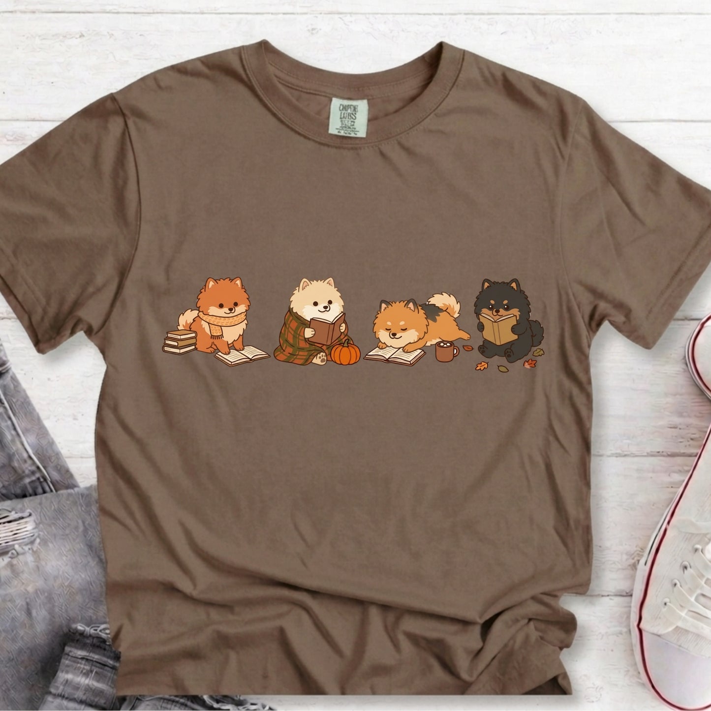 A brown short-sleeve crewneck t-shirt featuring a graphic of four cute cartoon dogs sitting and reading, styled on a white wood plank background with jeans and a sneaker partially visible.