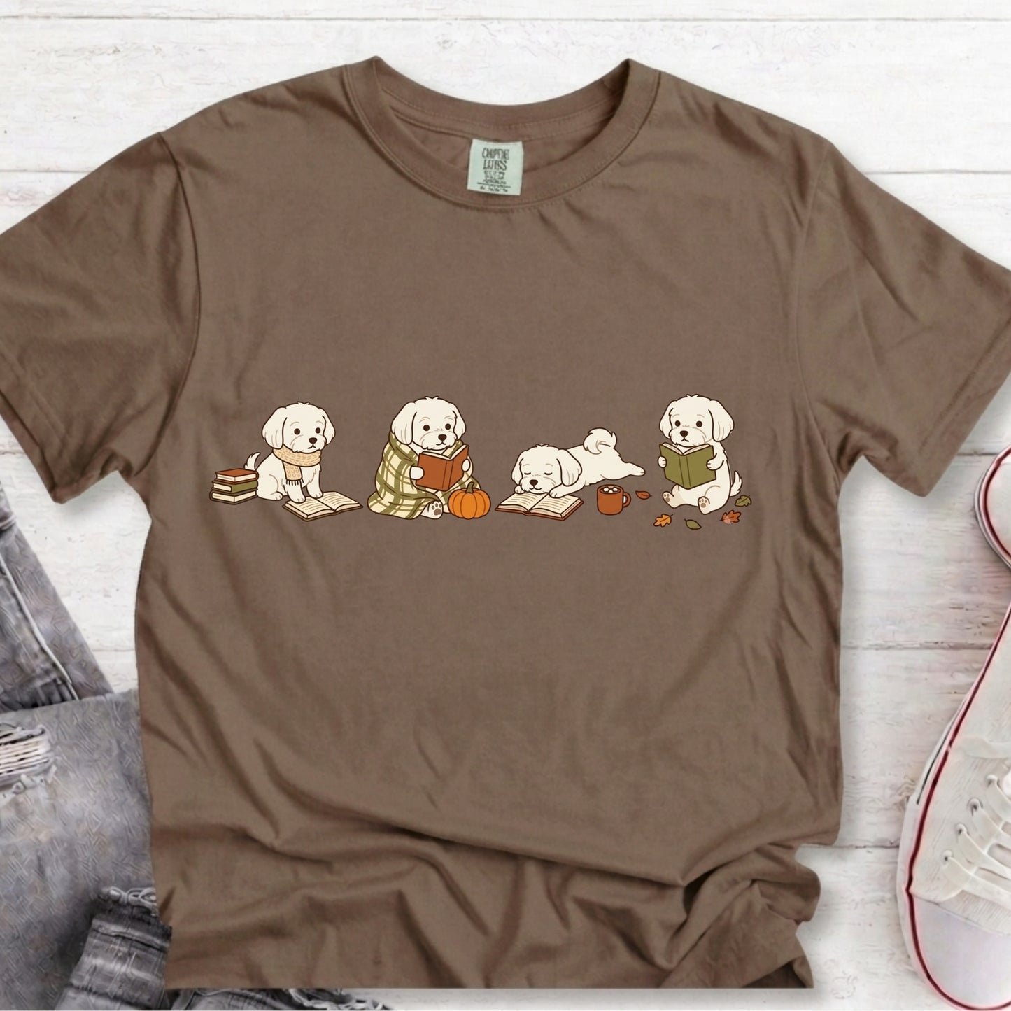 A brown short-sleeve crewneck t-shirt featuring a graphic of four cute white puppy dogs sitting and reading books, styled flat on a white wood background with jeans and a sneaker partially visible.