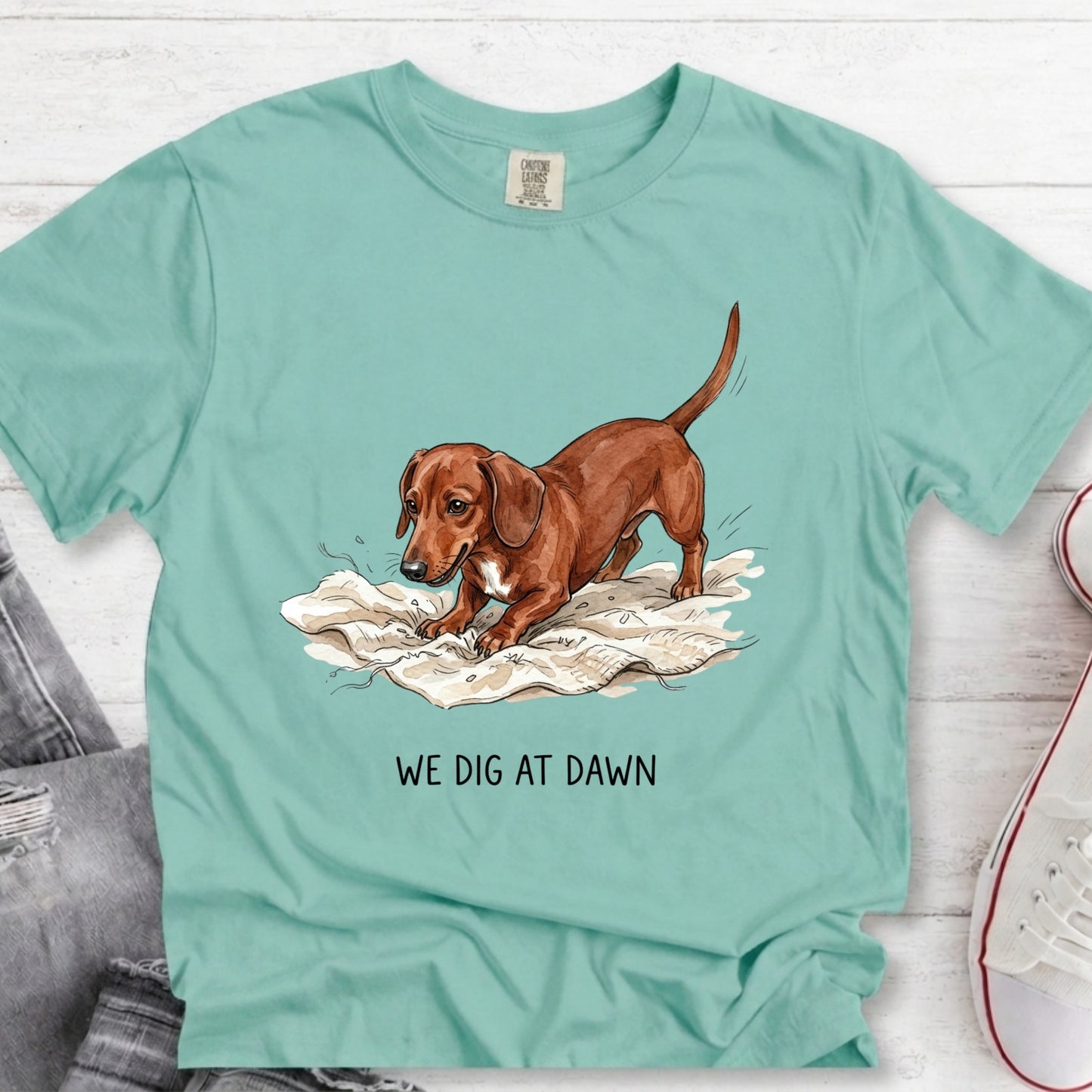 A mint green graphic t-shirt featuring a brown dachshund illustration digging in white newspaper, with the text 'WE DIG AT DAWN' printed below. The t-shirt is styled in a flat-lay on a white wood background with jeans and a sneaker partially visible.
