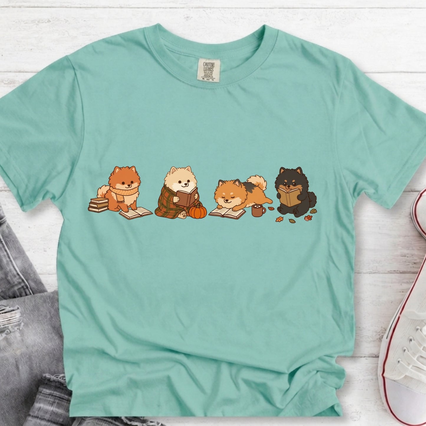 A mint green short-sleeve t-shirt laid flat on a white wood background, featuring a graphic of four cute cartoon cats reading books. The t-shirt is styled with a pair of gray jeans and a white sneaker in the corners.