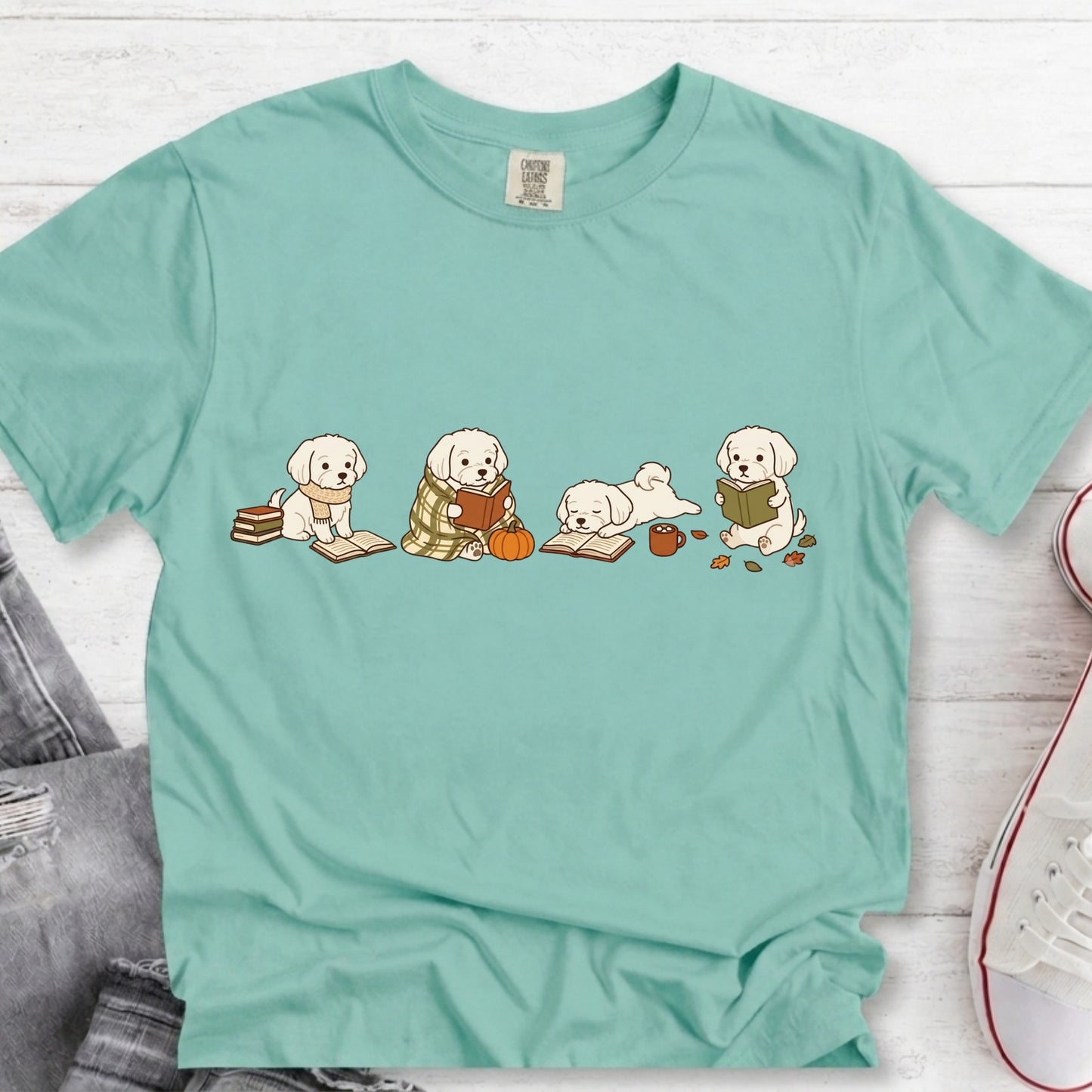 A mint green short-sleeve t-shirt with a graphic of four cute puppy characters reading books, laid flat on a white wood background with jeans and a sneaker partially visible.