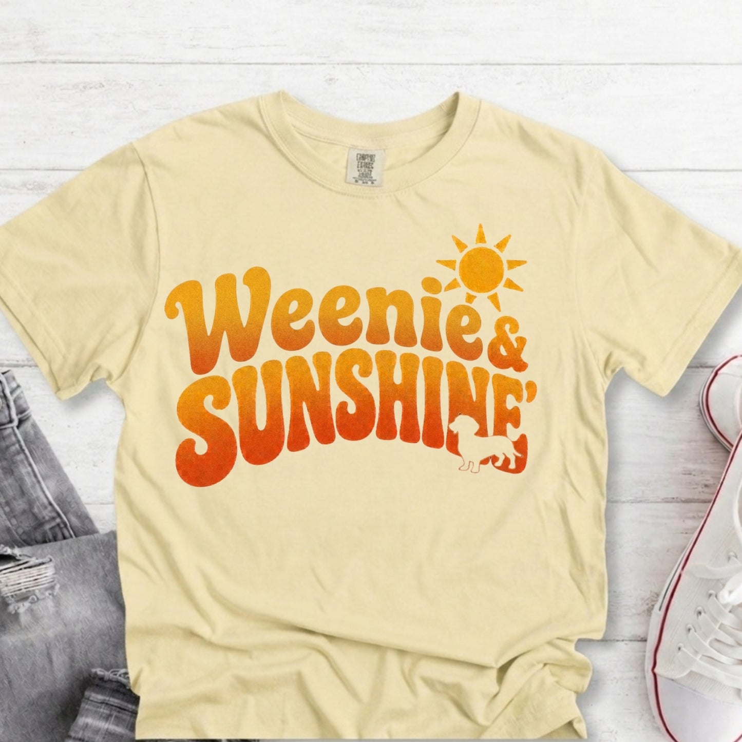 A pale yellow graphic T-shirt laid flat on a white wood surface, with distressed denim and a white sneaker partly visible at the edges. The shirt has a large retro-style orange and yellow gradient text reading 'Weenie & Sunshine' with a sun graphic and small bear silhouette.