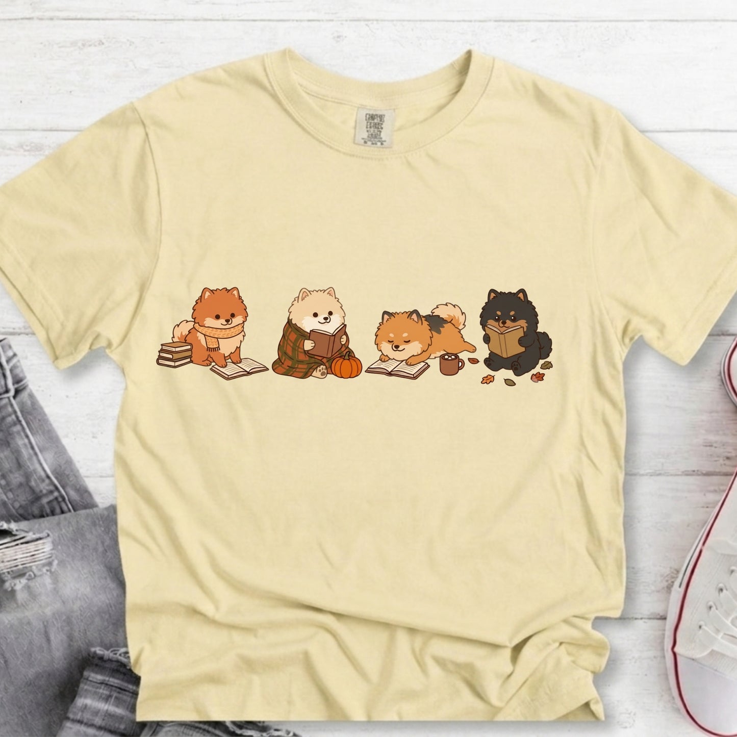 A pale yellow short-sleeve t-shirt with a graphic of four cute cartoon dogs reading books, laid flat on a white wood background with jeans and a sneaker partially visible.