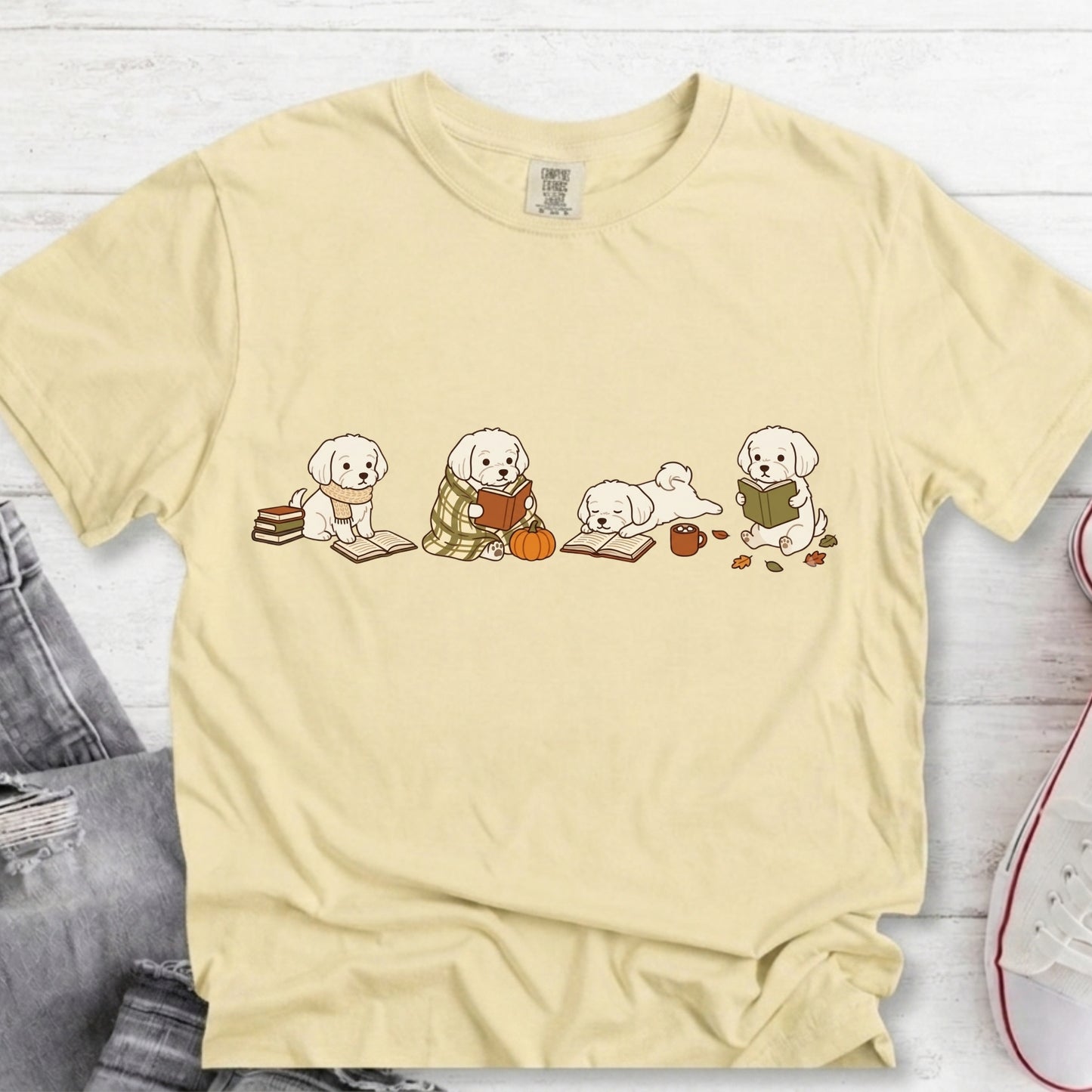 A pale yellow short-sleeve t-shirt with a graphic of four white puppies reading books, styled in a flat-lay on a white wood background with jeans and a sneaker partially visible.