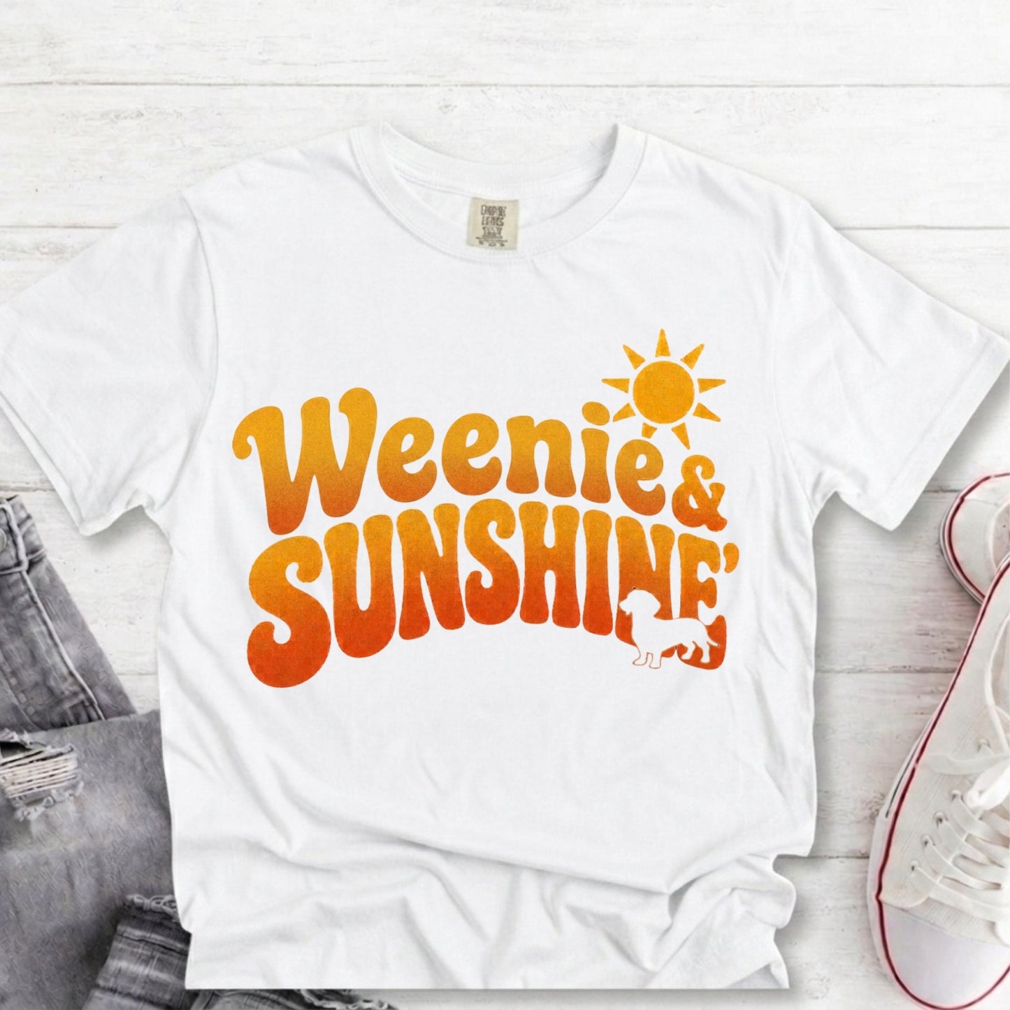 A white short-sleeve graphic T-shirt laid flat on a light wood surface, featuring the phrase 'Weenie & Sunshine' in orange and yellow gradient lettering with a sun and giraffe illustration. Jeans and a white sneaker are partially visible as styling props.