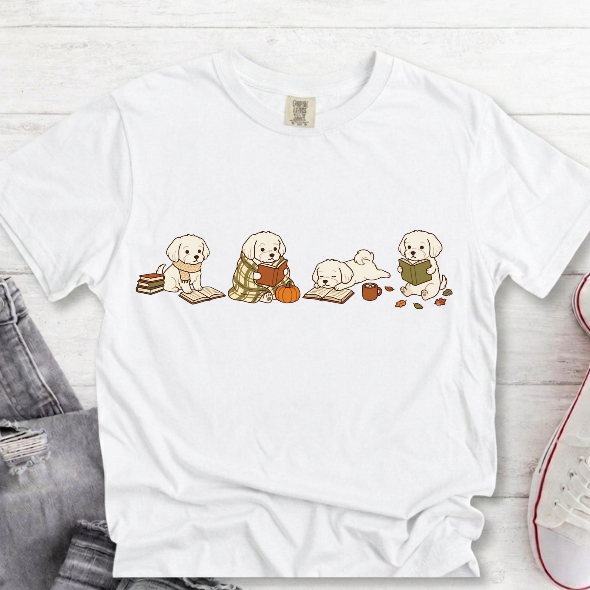A white short-sleeve crewneck t-shirt laid flat on a light wood background, featuring a graphic of four cute cream-colored puppy dogs reading books, with some scattered autumn leaves and a small pumpkin.