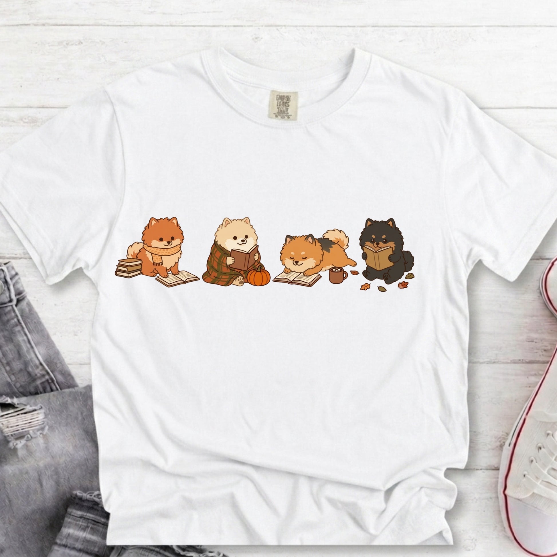 A white short-sleeve crewneck T-shirt laid flat on a light wood surface, featuring a horizontal cartoon print of four fluffy cats reading books. The shirt is styled with a pair of gray jeans on the left and a white sneaker on the right.