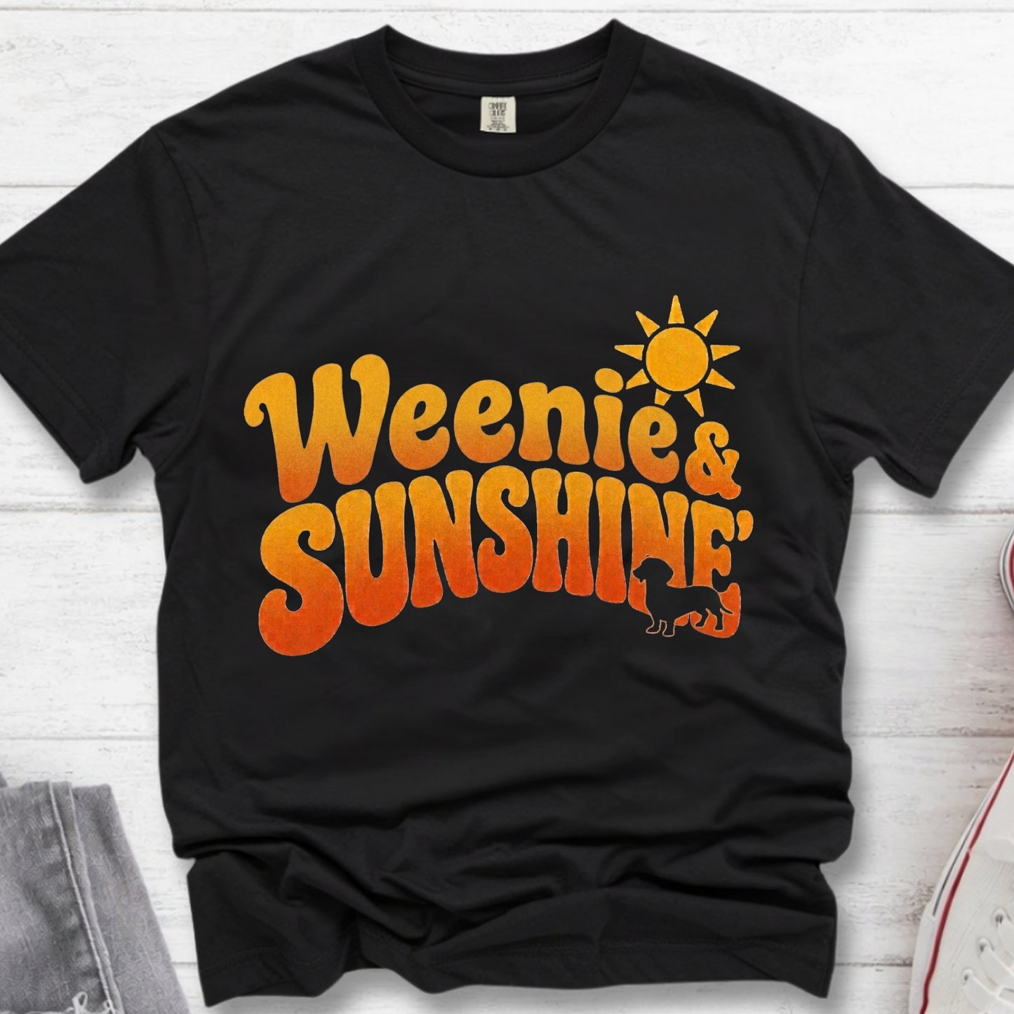 A black short-sleeve t-shirt laid flat on a white wood background, featuring a retro-style orange and yellow gradient graphic that reads 'Weenie & Sunshine' with a sun and silhouette.