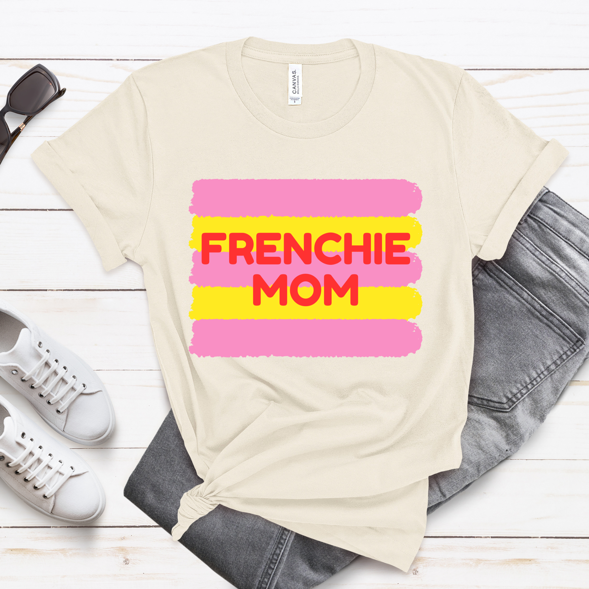 Beige t-shirt with 'FRENCHIE MOM' text on a wooden surface with shoes and jeans.