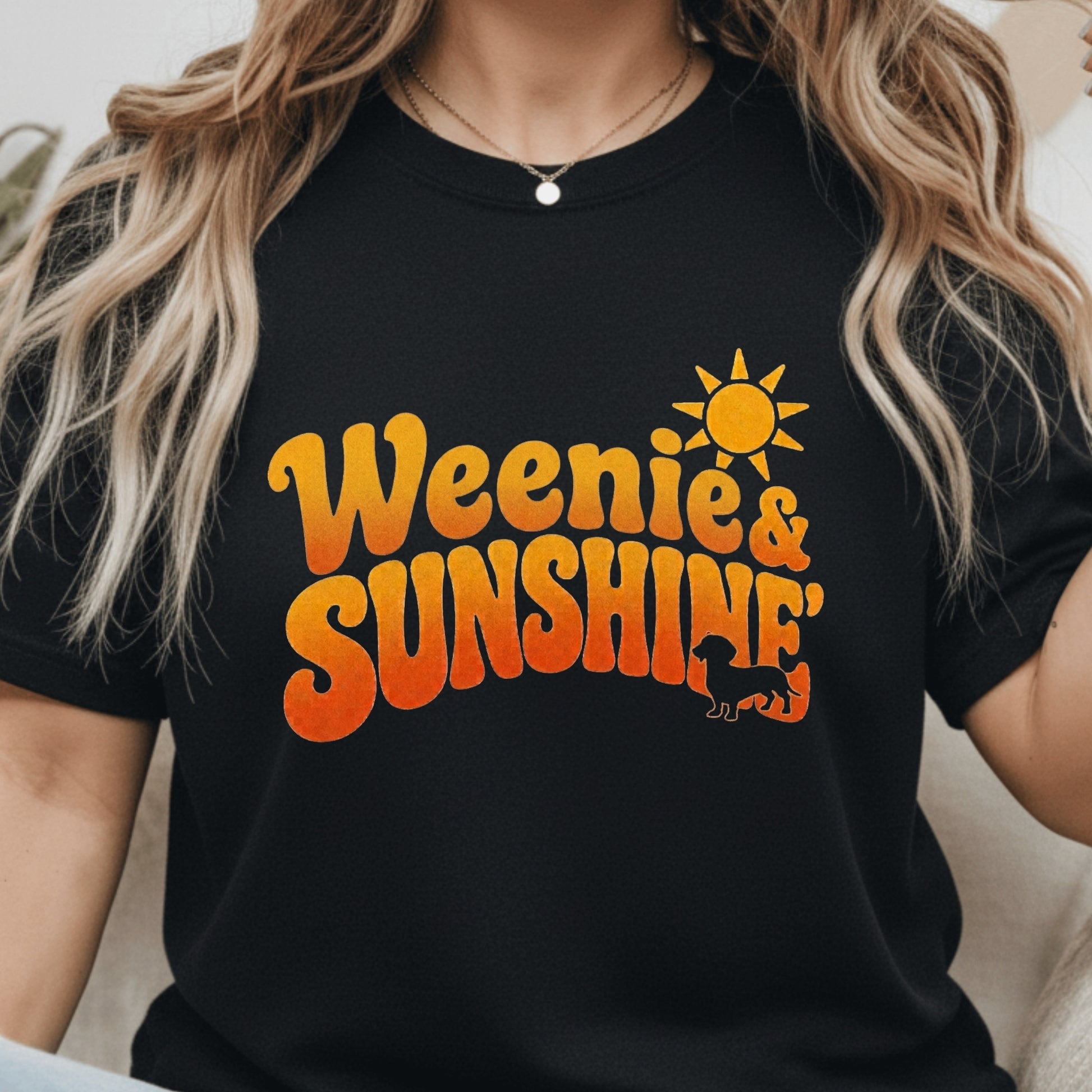 A woman wearing a black short-sleeve t-shirt with a retro-style orange and yellow graphic that reads 'Weenie & Sunshine' with a sun and cat silhouette.