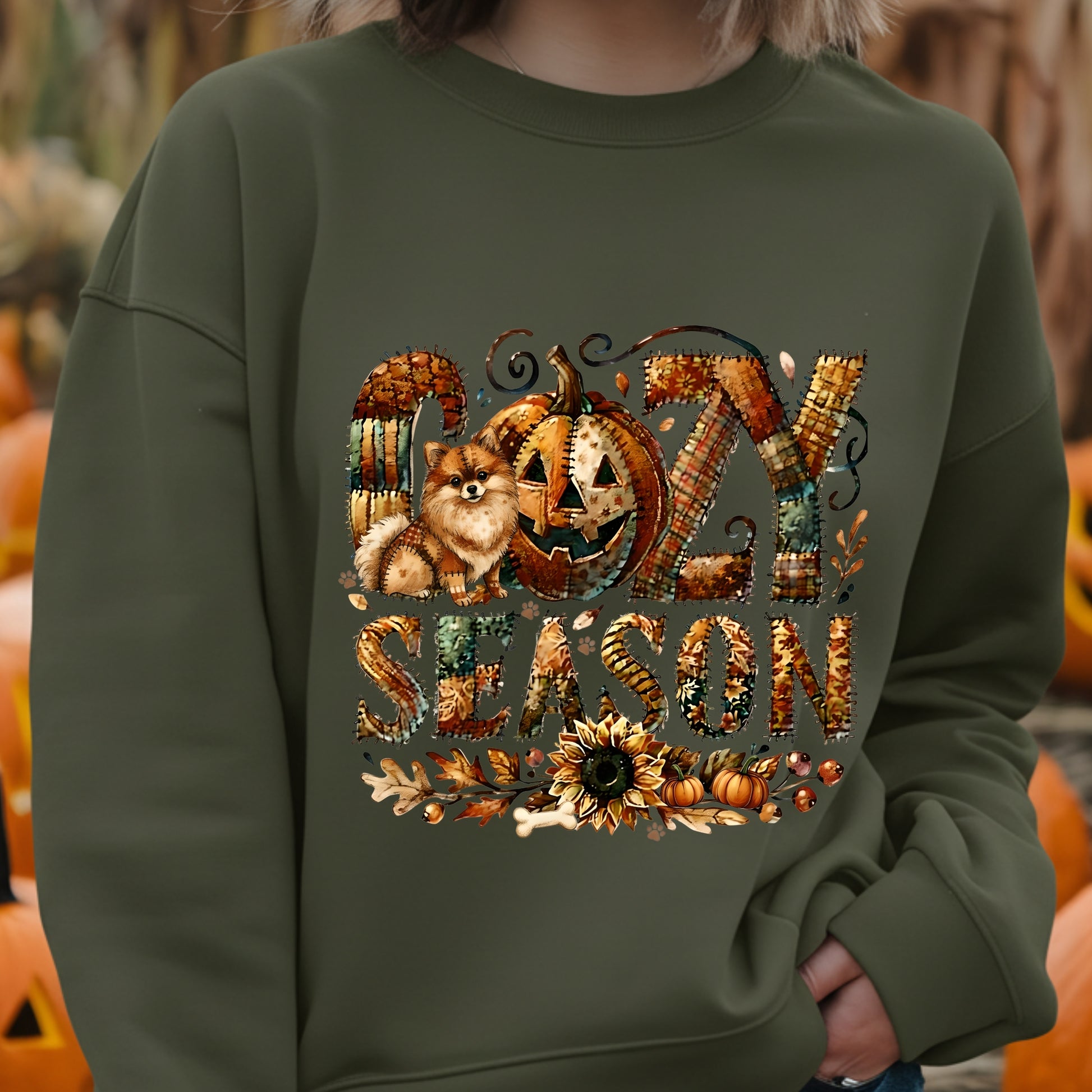 Green sweatshirt with 'Cozy Season' design featuring pumpkins and a dog, set against a blurred autumn background.