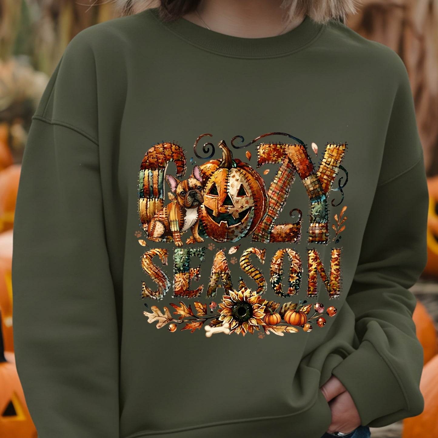 Person wearing an military green sweatshirt with ' cozy season' design featuring pumpkins and leaves.