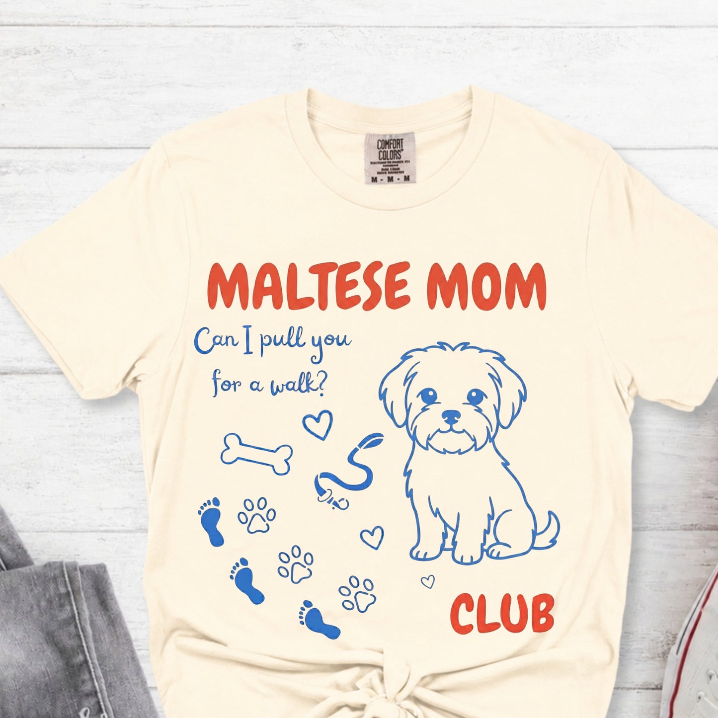Beige t-shirt with Maltese dog graphic and text on a light wooden surface