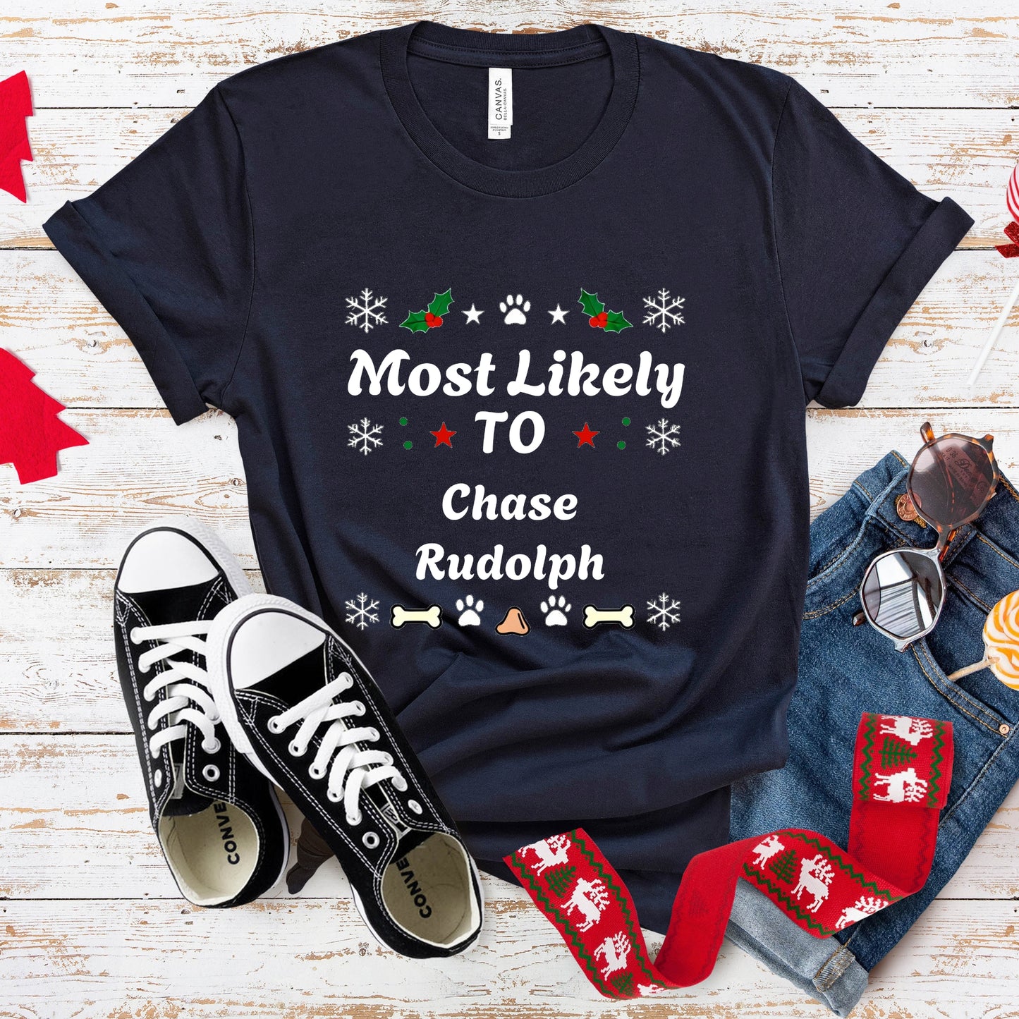 Navy blue t-shirt with 'Most Likely To Chase Rudolph' text, black sneakers, red socks, and sunglasses on a wooden surface.