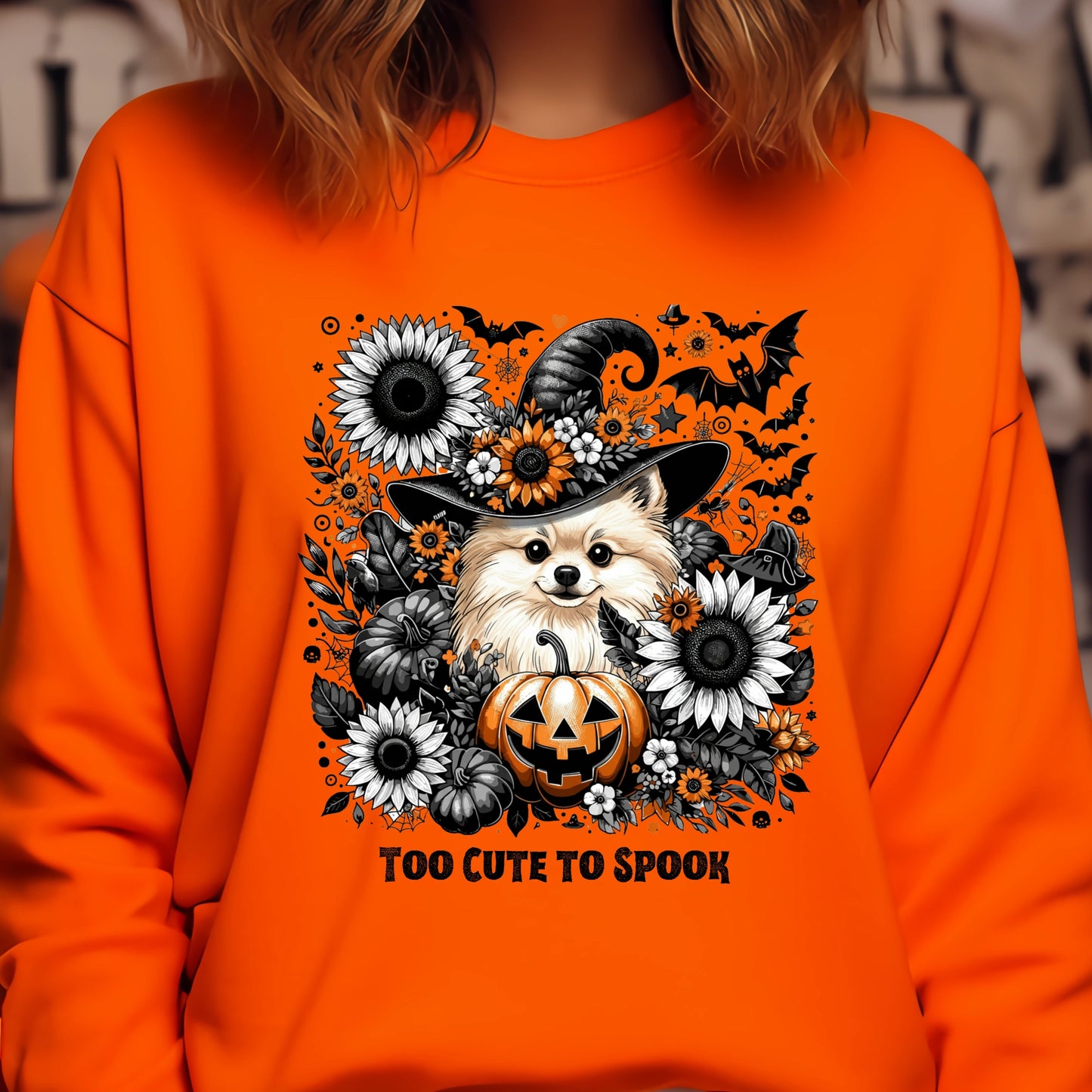 Orange sweatshirt with Halloween-themed design featuring a dog in a witch hat and pumpkin, surrounded by flowers and text 'Too Cute to Spook'.