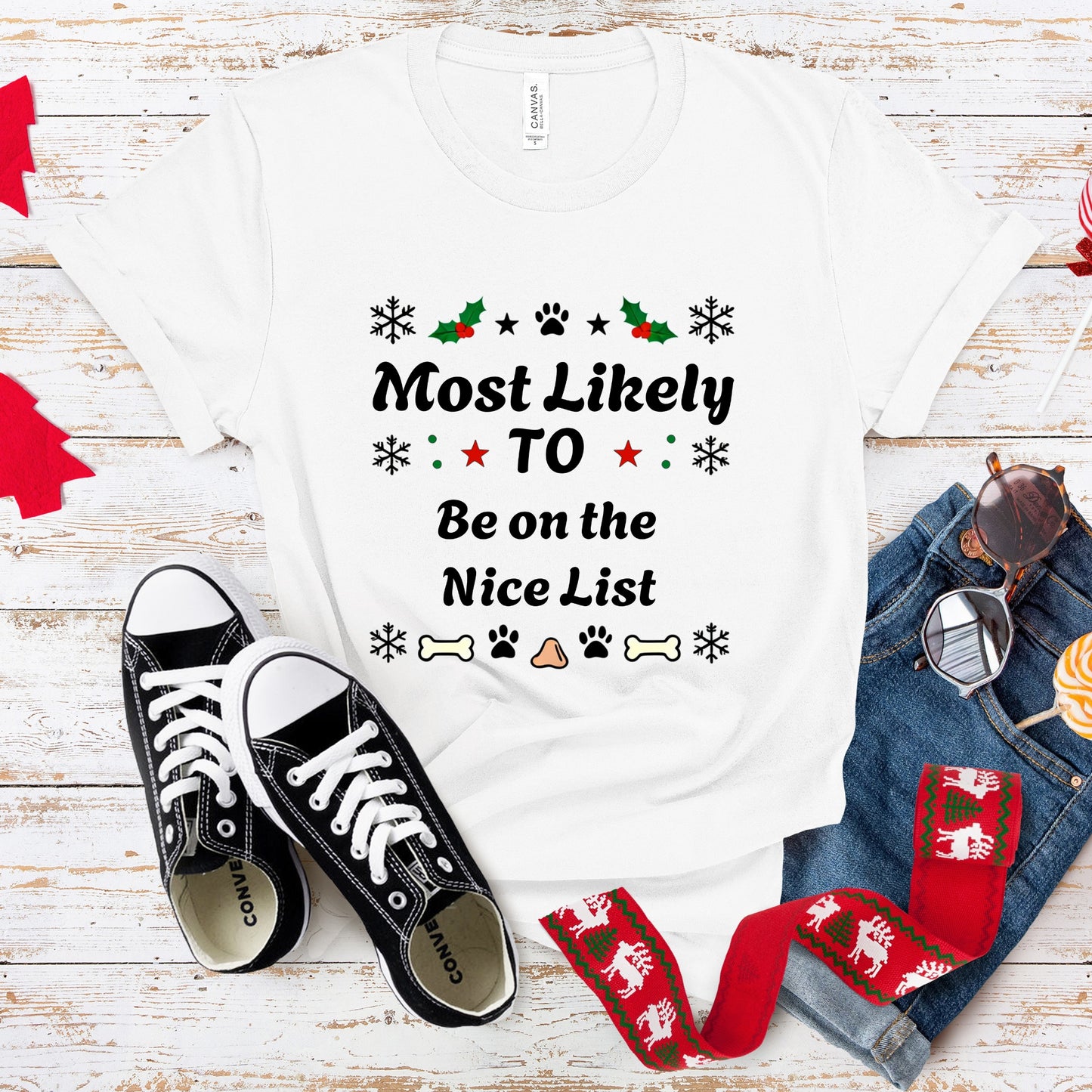 White t-shirt with Christmas-themed text, black sneakers, red and green scarf, and jeans on a wooden surface.