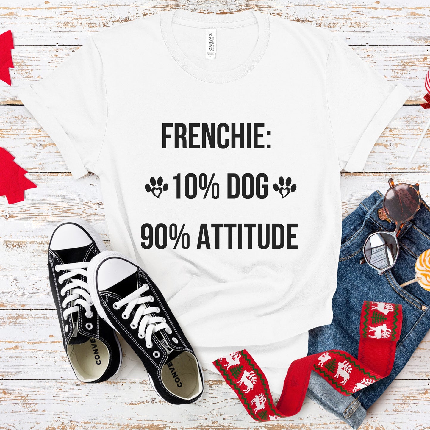 White t-shirt with 'FRENCHIE: 10% DOG 90% ATTITUDE' text, black sneakers, red and green patterned bag, and jeans on a wooden surface.