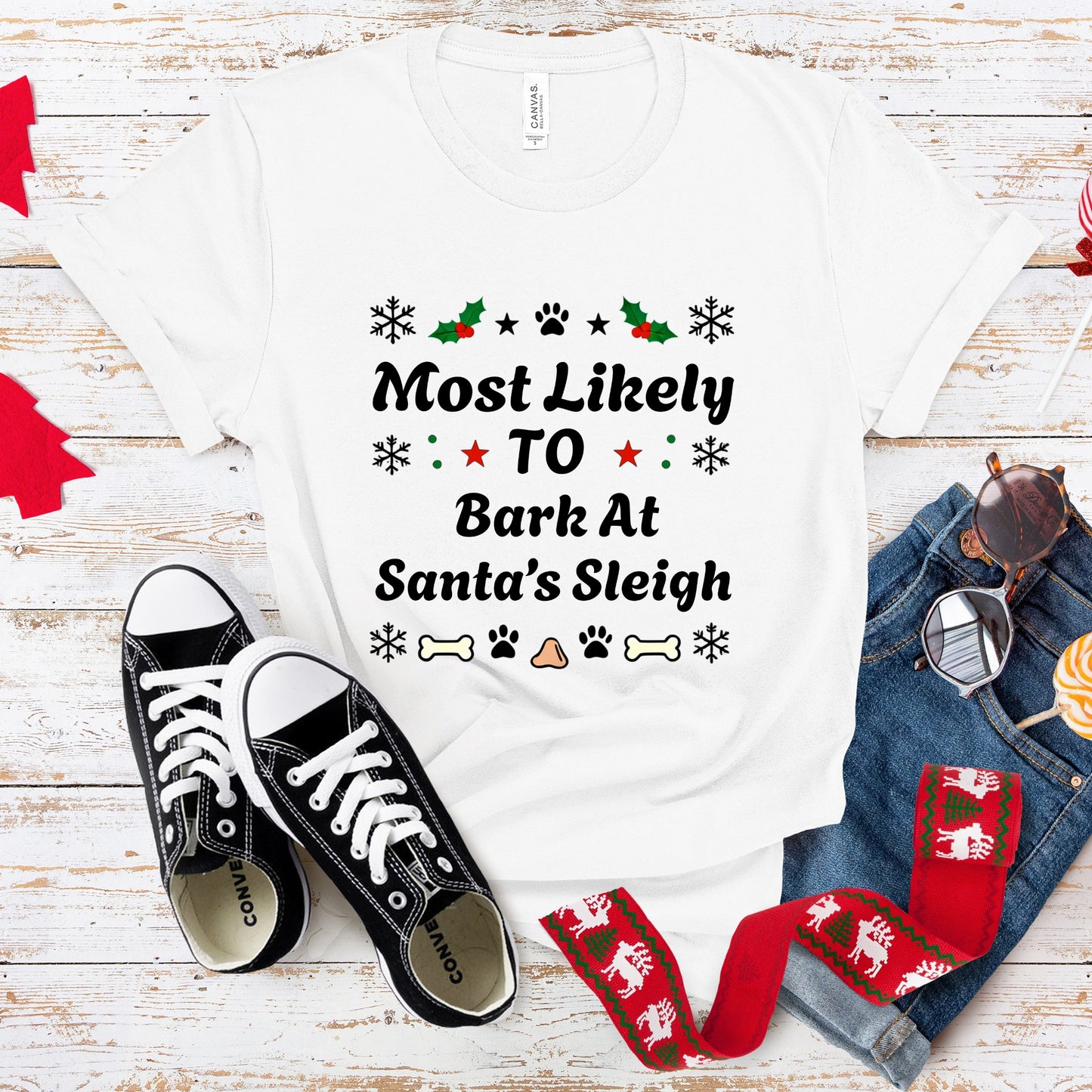 White t-shirt with Christmas-themed text, black sneakers, and festive accessories on a wooden surface.