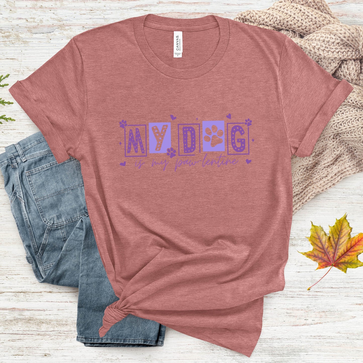 Mauve t-shirt with 'My dog is my first teacher' text on a wooden surface with jeans and leaves.