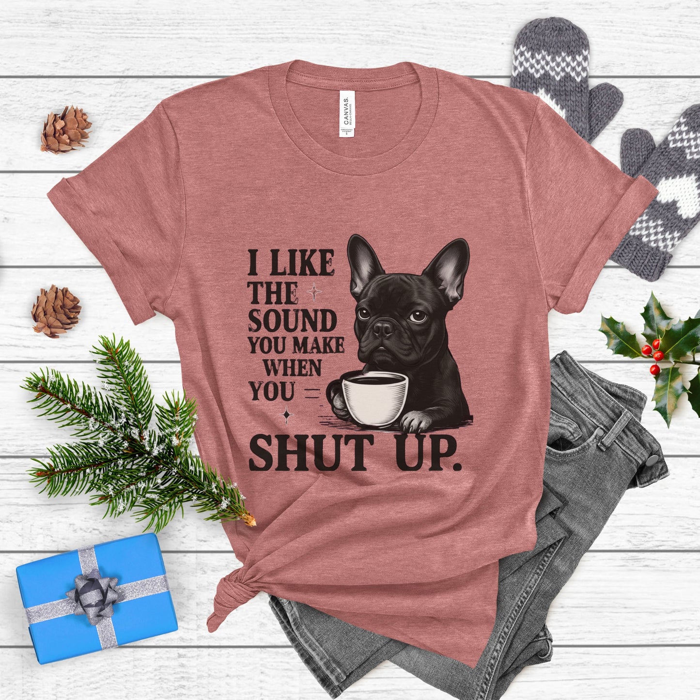 Mauve t-shirt with a graphic of a dog holding a coffee cup and text, placed on a wooden surface with festive decorations.
