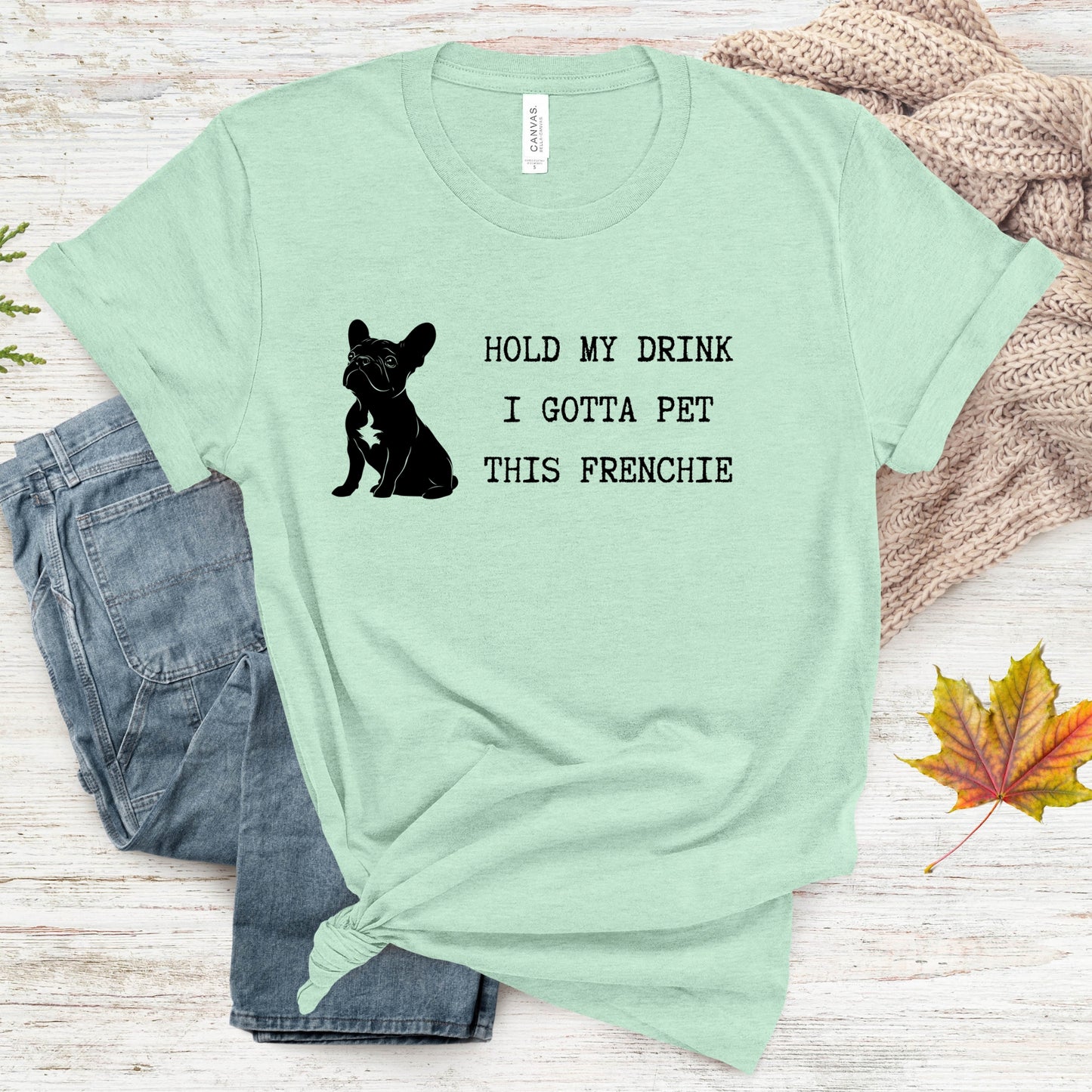 Mint green t-shirt with black text and dog graphic on a wooden surface with jeans and leaves.