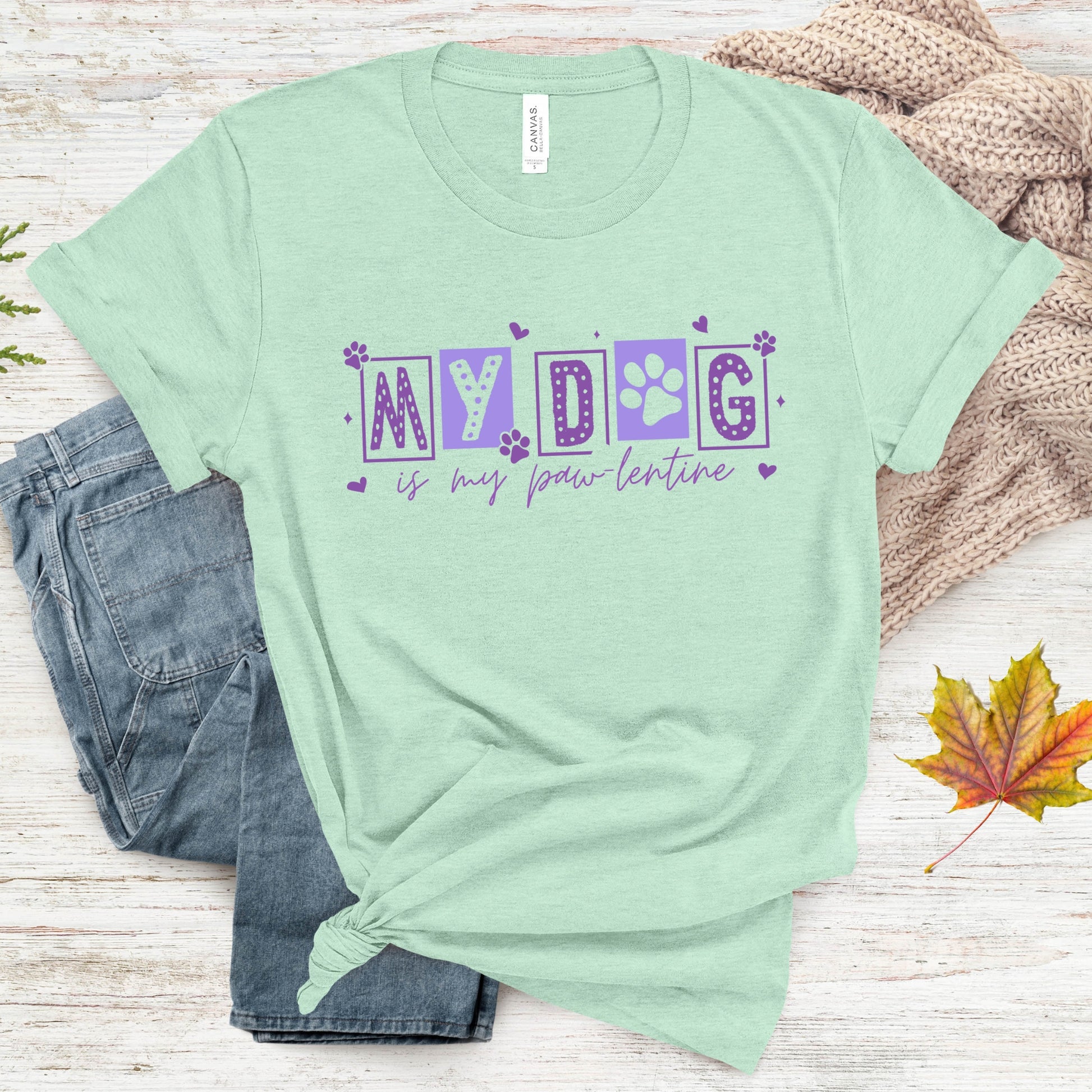 Mint green t-shirt with 'My dog is my paw-lentine' text on a wooden surface with jeans and leaves.