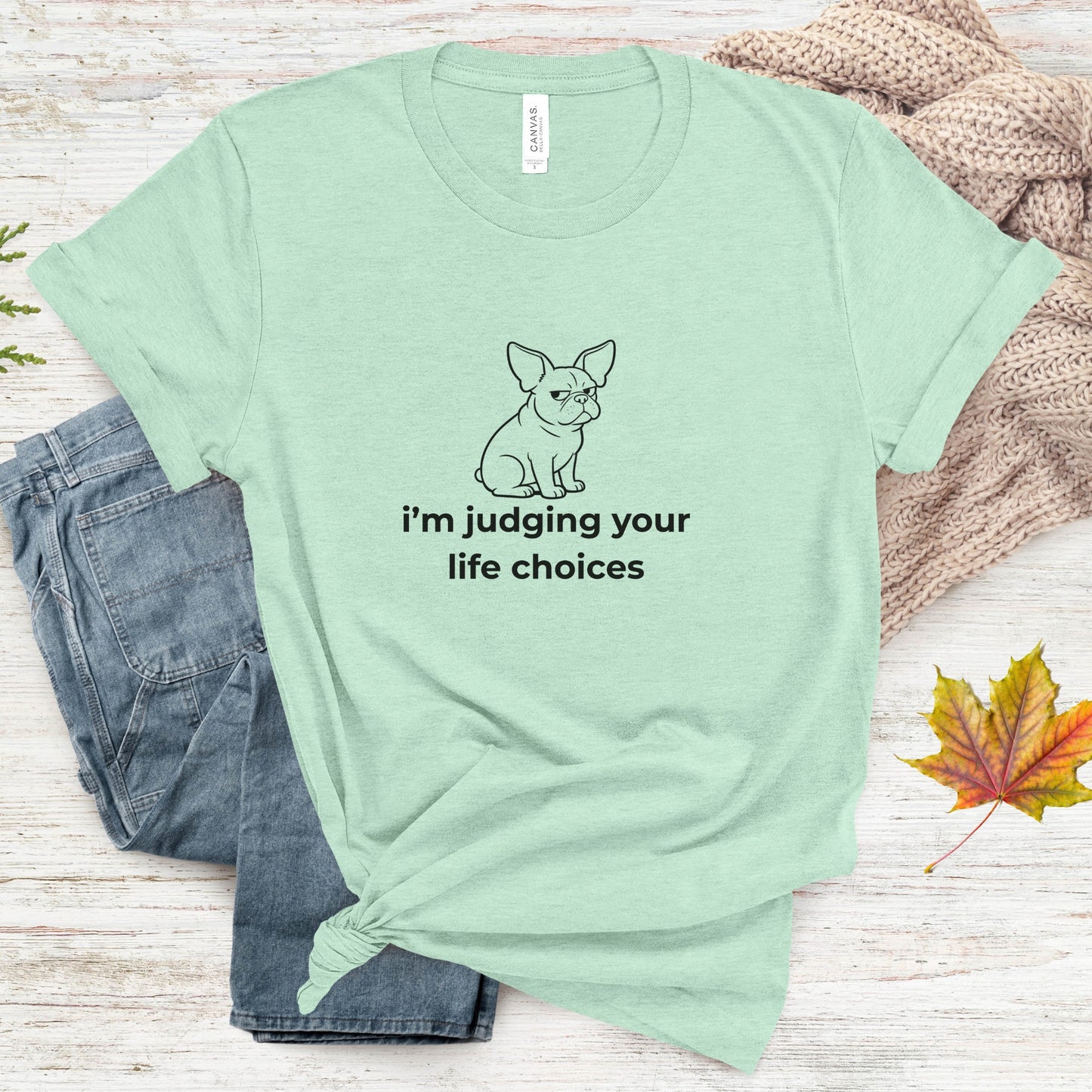 Mint green t-shirt with a dog graphic and text, placed on a wooden surface with jeans and leaves.