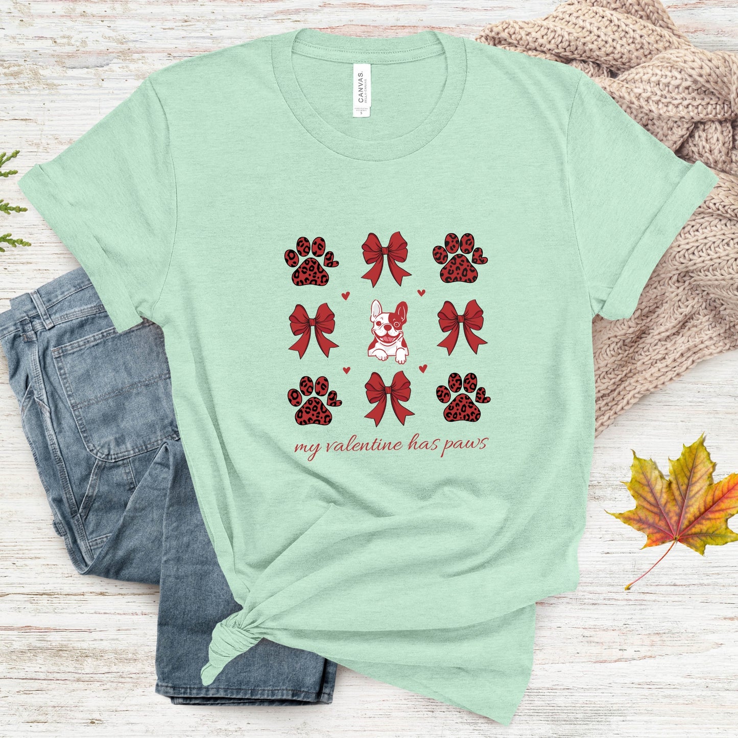 Mint green t-shirt with red paw and bow designs and text 'my valentine has paws' on a wooden surface.