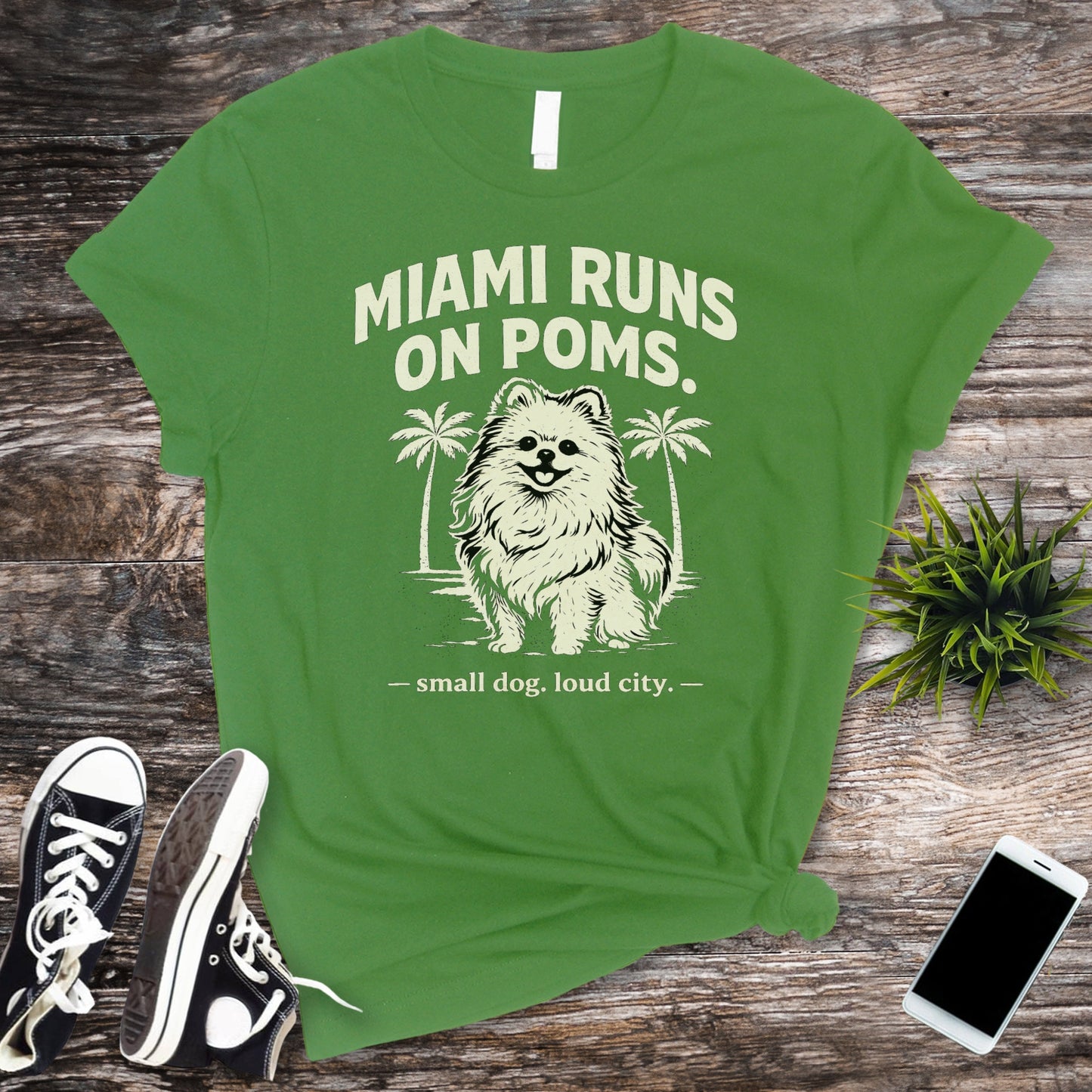 Green t-shirt with a dog graphic and text on a wooden surface with shoes and a phone.