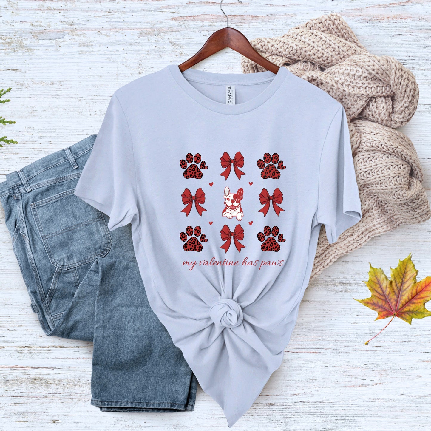 Light blue t-shirt with red paw and bow design, paired with jeans and a sweater on a wooden surface.