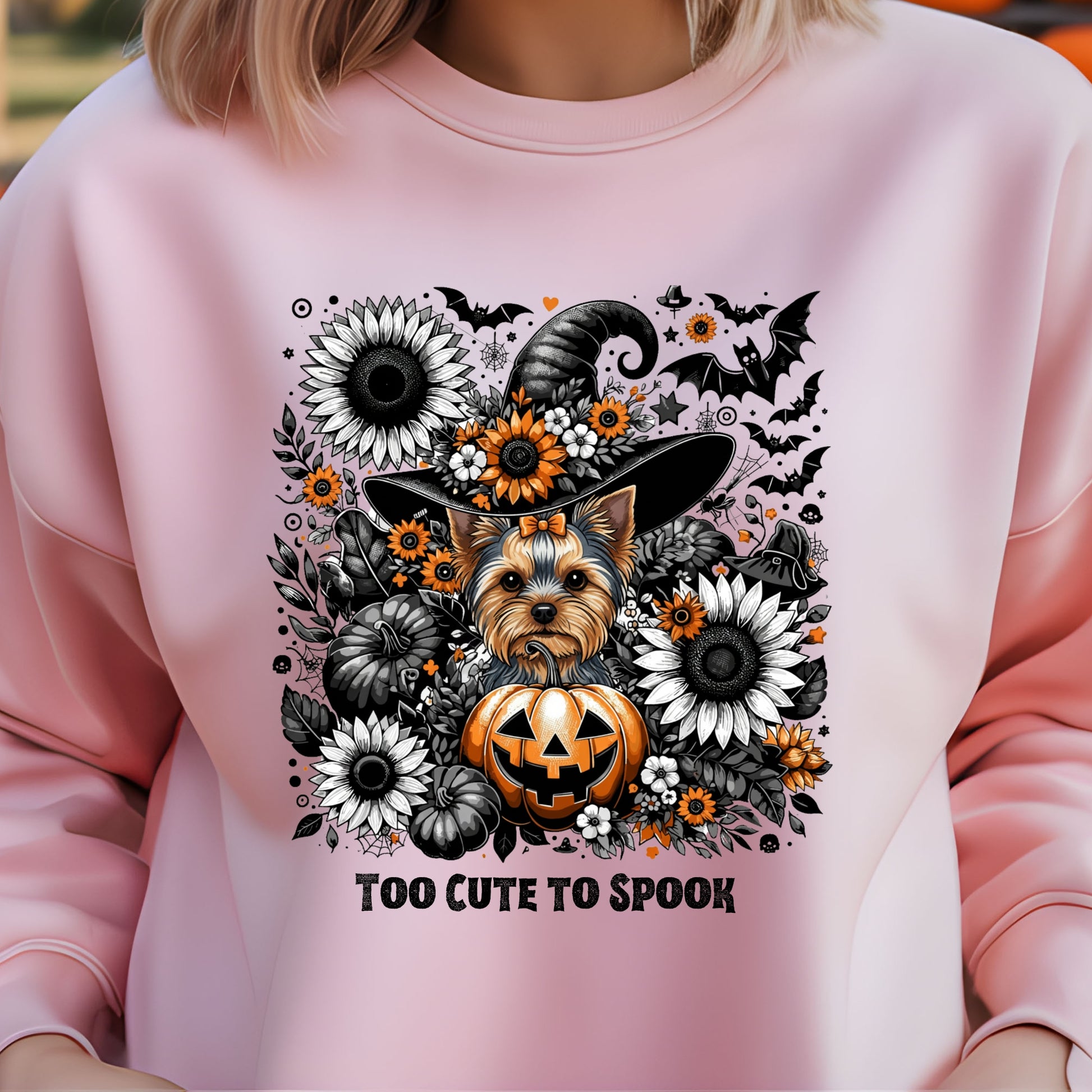 Pink sweatshirt with a Halloween-themed design featuring a dog, pumpkins, and flowers on a blurred background.
