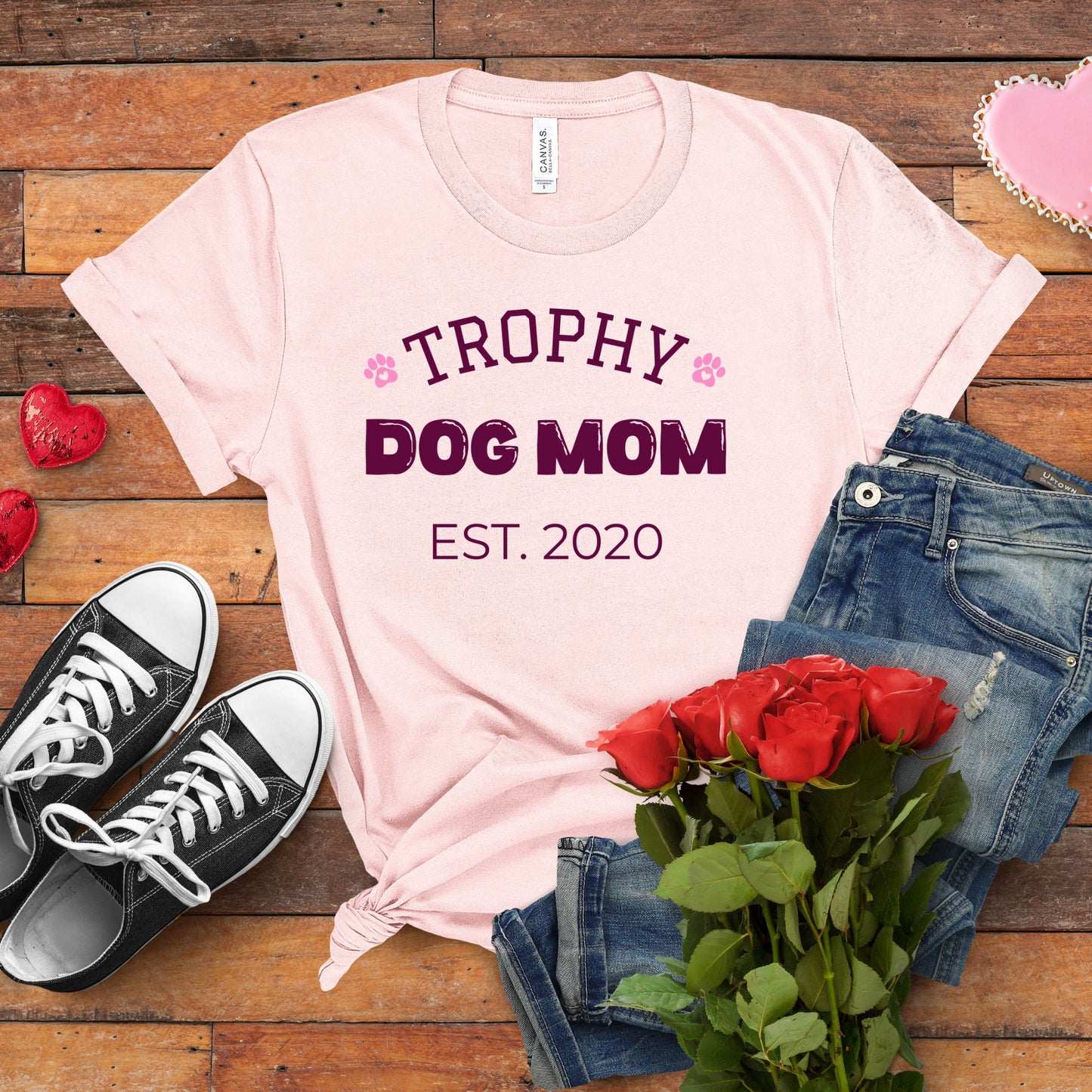 Pink t-shirt with 'Trophy Dog Mom Est. 2020' text, black sneakers, red roses, and jeans on a wooden surface.