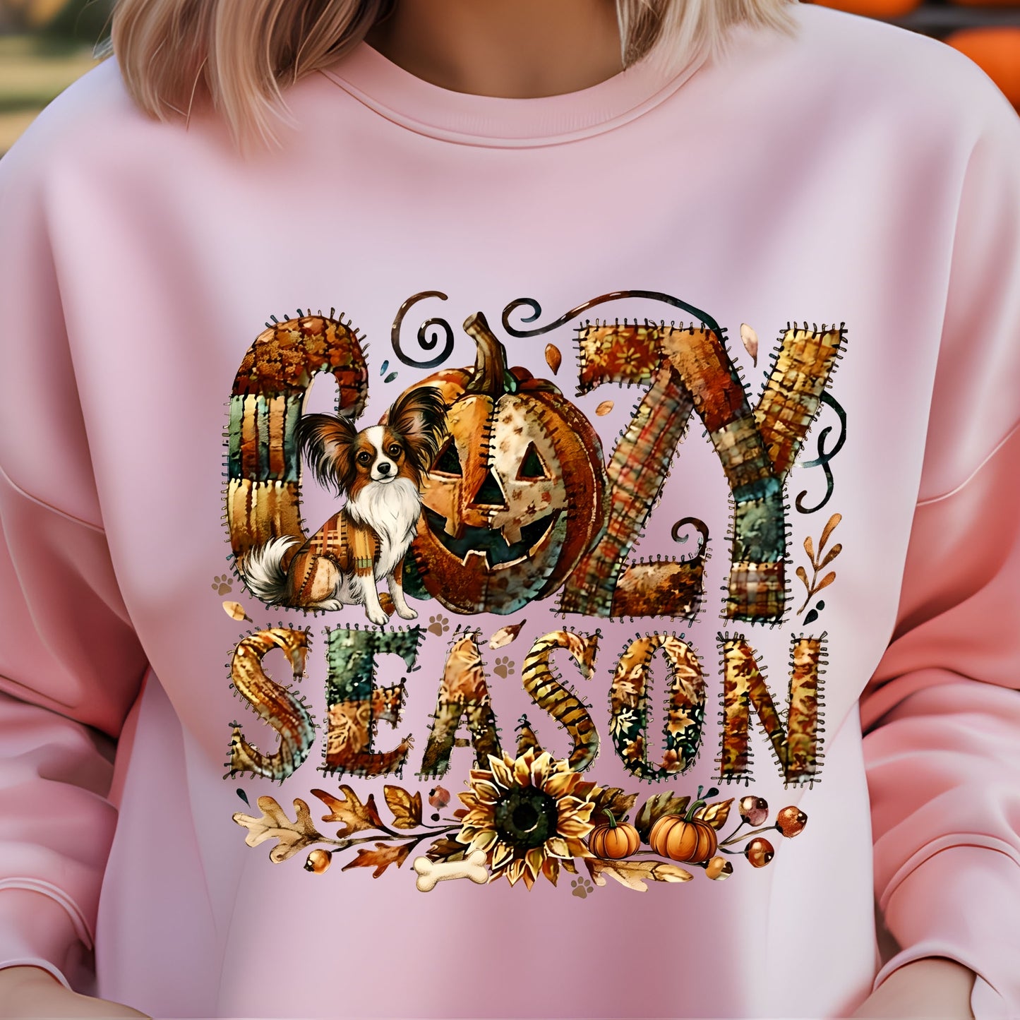 Pink sweatshirt with Halloween-themed design featuring a pumpkin, dog, and text on a blurred background.