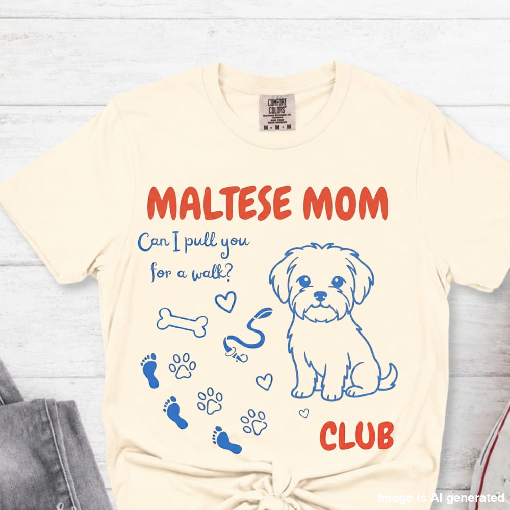 Beige t-shirt with Maltese dog graphic and text on a light wooden surface