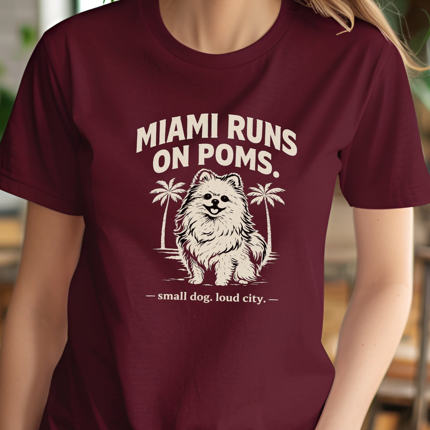 Maroon t-shirt with a graphic of a dog and text 'MIAMI RUNS ON POMS' on a blurred background