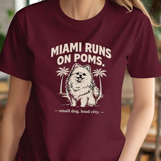 Maroon t-shirt with a graphic of a dog and text 'MIAMI RUNS ON POMS' on a blurred background
